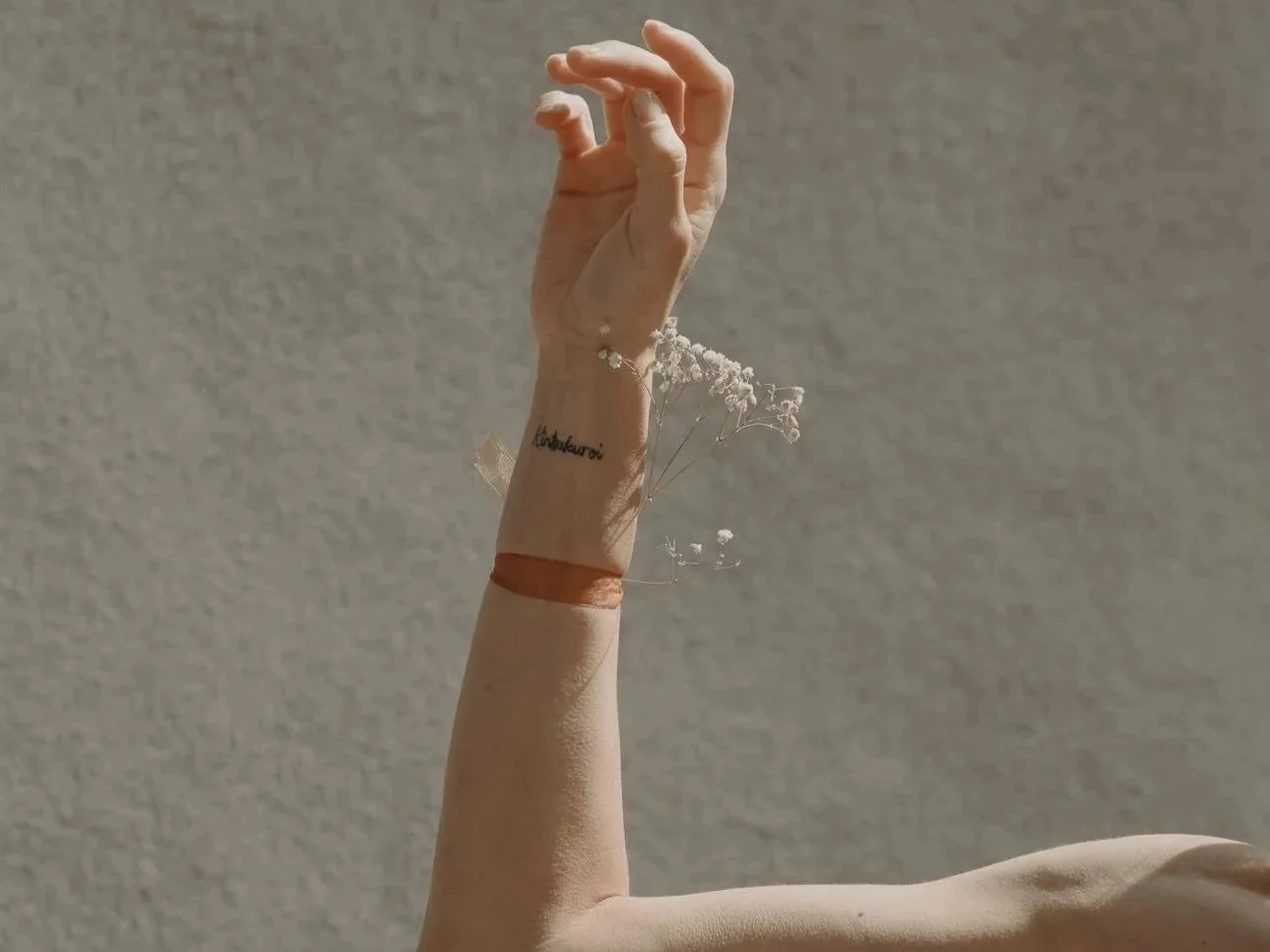 Polso e avambraccio di una persona con un tatuaggio e un braccialetto di cuoio, con fiori secchi come decorazione, contro uno sfondo di muro grigio