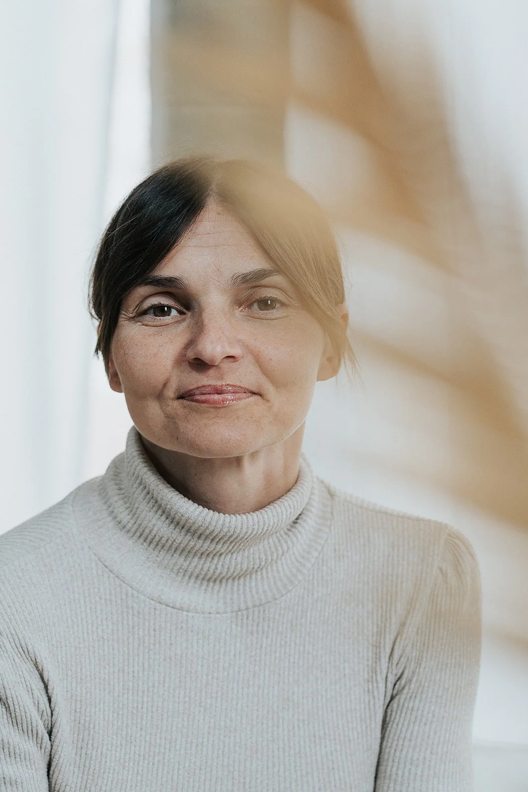 Interno con vista su una donna con capelli corti castani, carnagione chiara, indossa un maglione a collo alto beige, sfondo neutro, luce naturale.