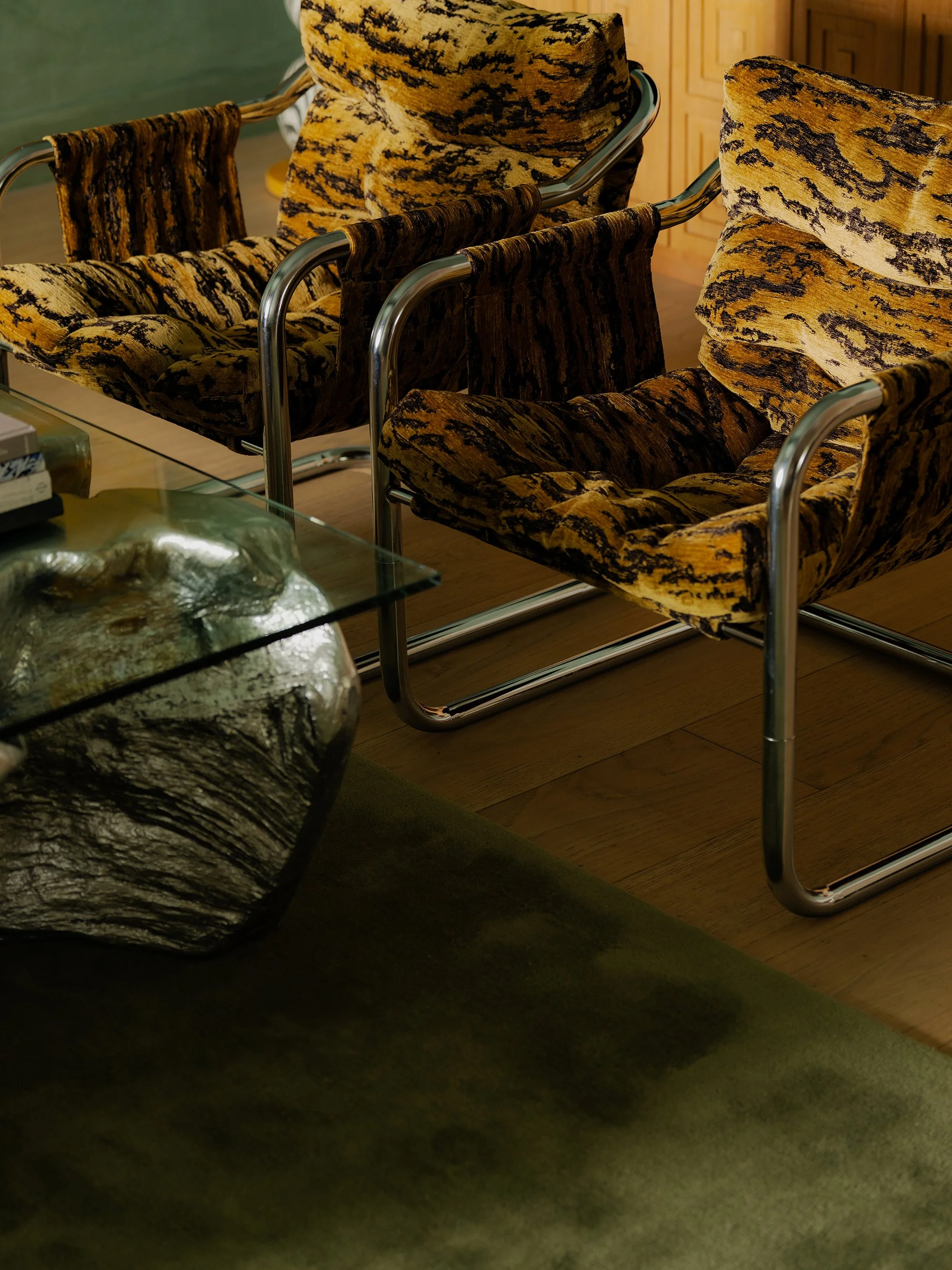 A living room scene with patterned armchairs, a glass coffee table, a black textured sofa, and a wooden wall with built-in cabinets.