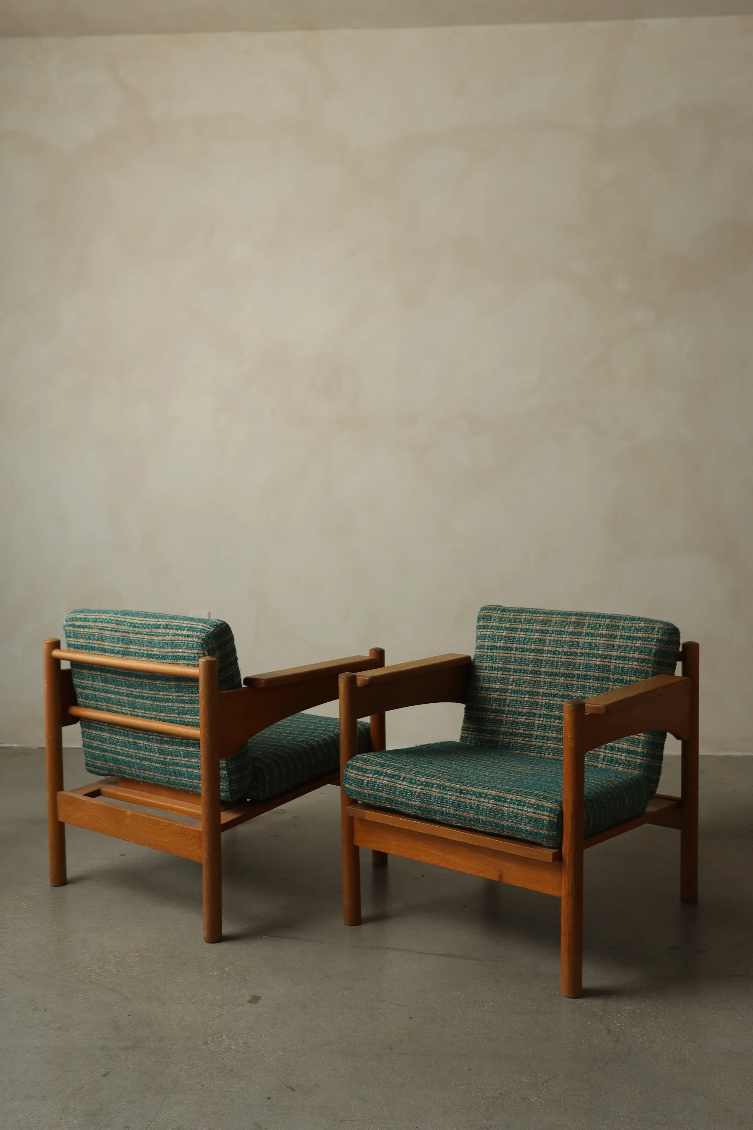 Two wooden chairs with green and blue striped cushions placed against a plain beige wall on a gray concrete floor.
