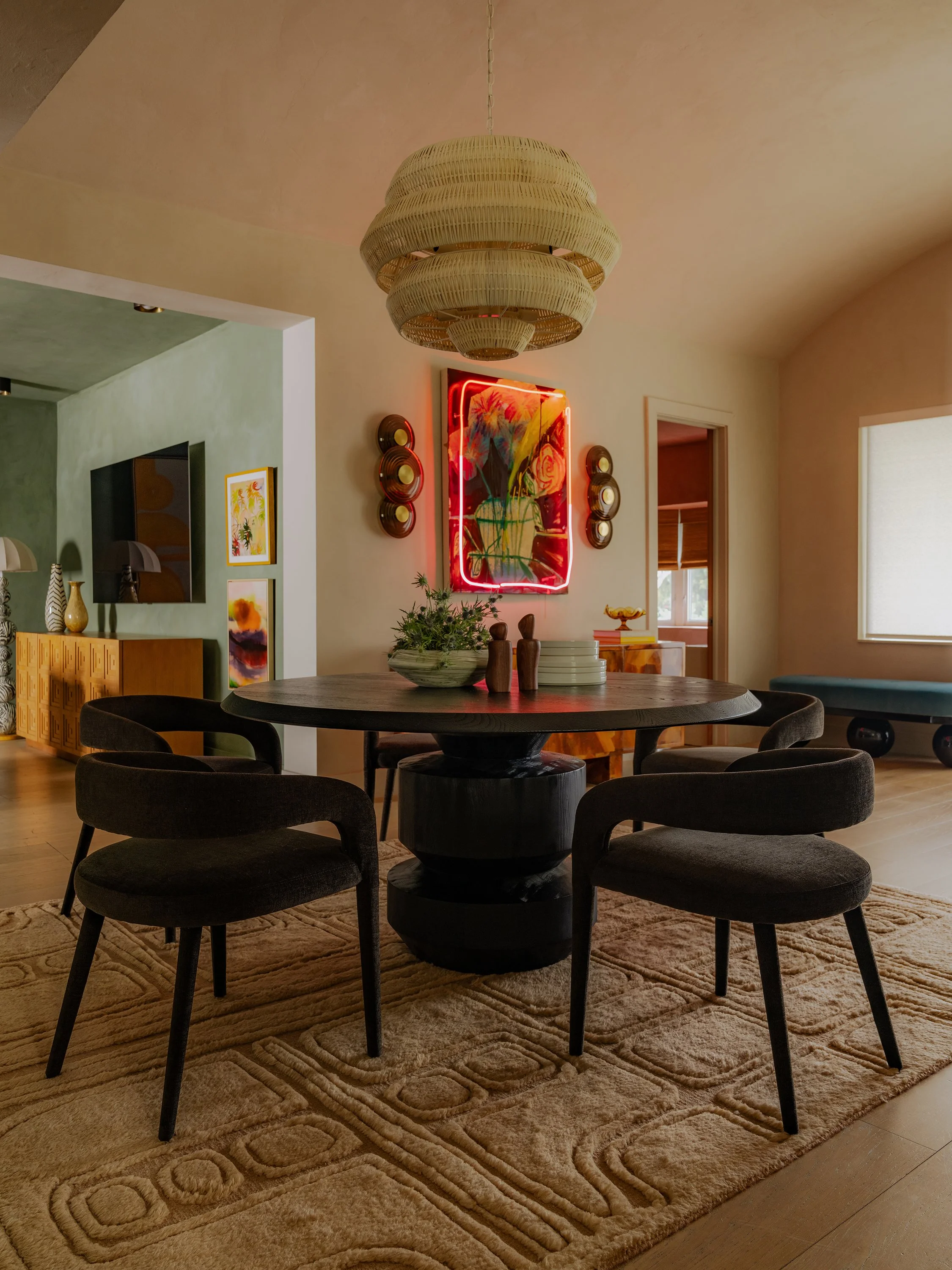 A round dining table with six black chairs on a beige patterned rug in a living room. A decorative bowl with green foliage and small wooden sculptures are on the table. The room features a large woven pendant light, colorful artwork with neon lighting on the wall, and a mix of modern and vintage decor.