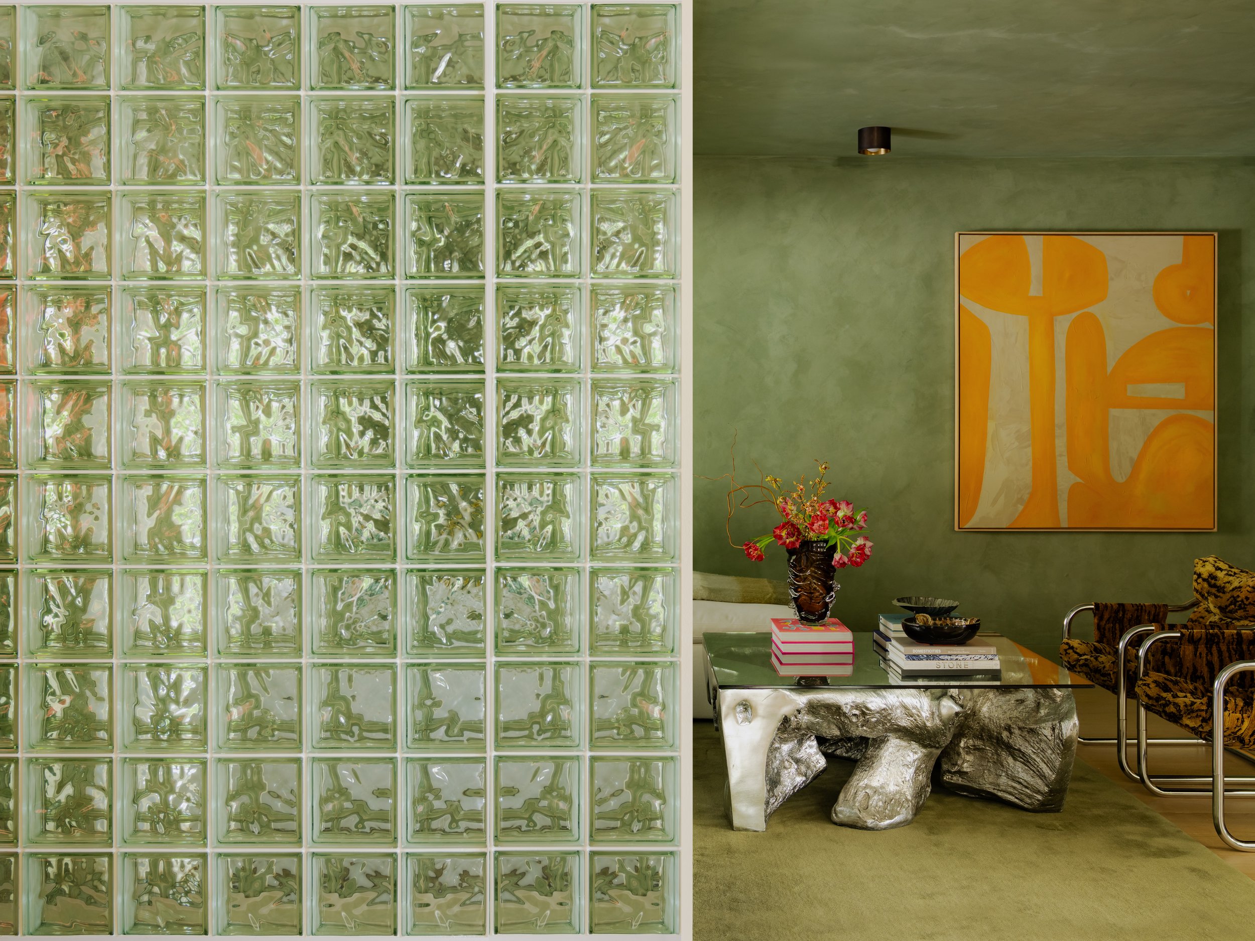 Interior view of a modern living space featuring a large glass block wall, a green wall with a painting, a side table with books and a flower vase, and chairs with patterned upholstery.