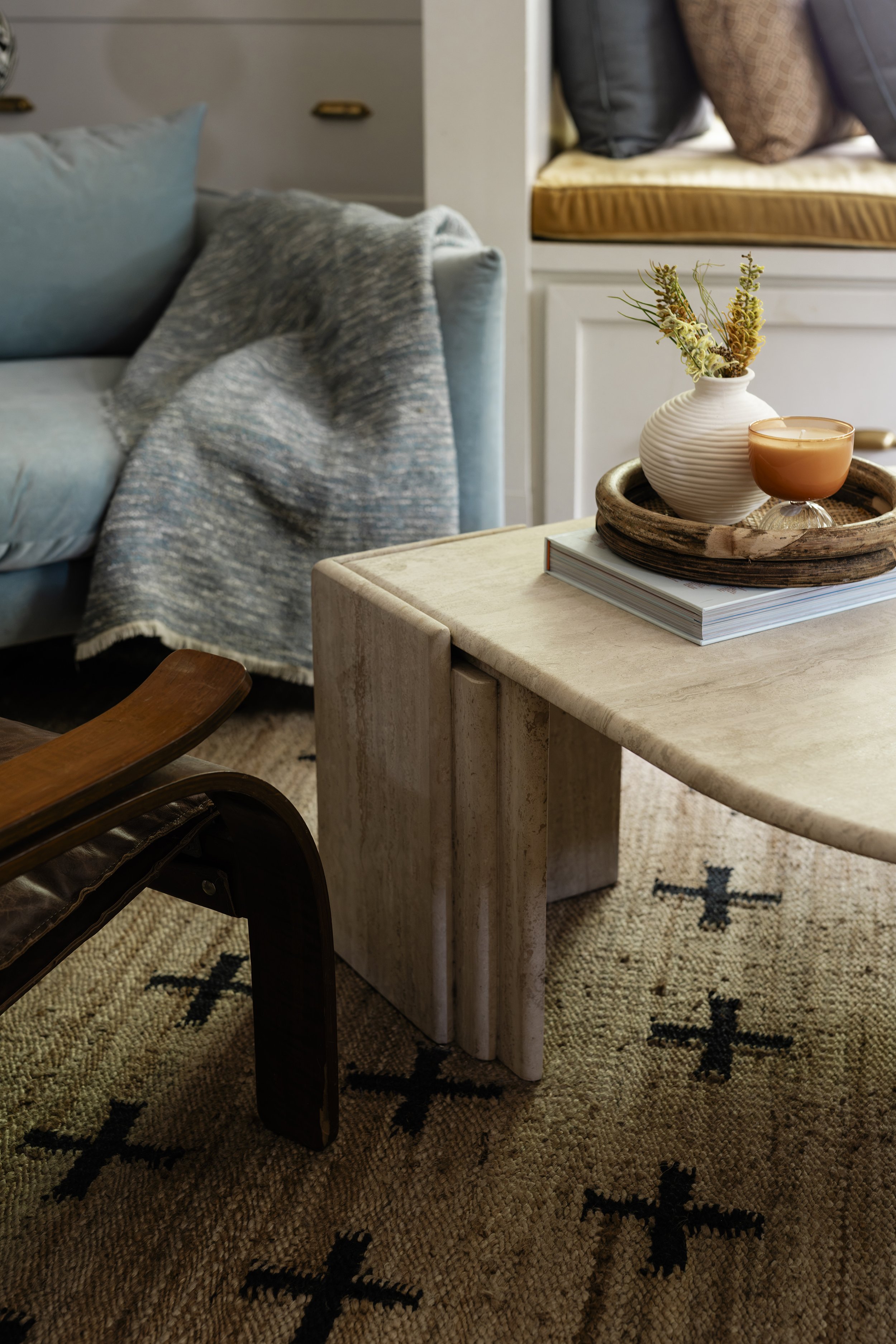 Close-up of a cozy living room corner with a beige side table holding a white vase with dried flowers, a candle, and a book. There is a blue sofa with a textured throw blanket and pillows, a patterned rug on the floor, and a built-in window seat with cushions in the background.