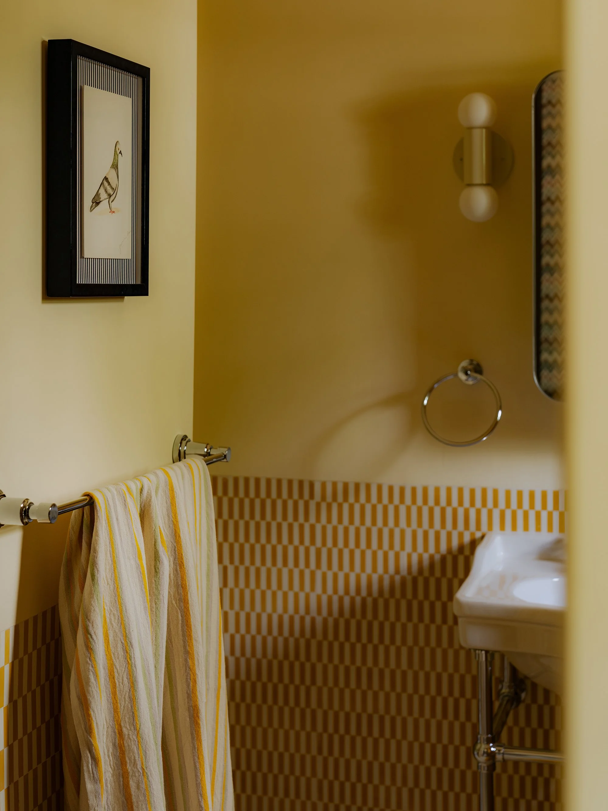 A bathroom interior featuring a framed picture of a pigeon, a yellow wall, a towel rack with yellow and white towels, a wall-mounted light fixture, a small white sink, and a decorative tile pattern on the lower part of the wall.