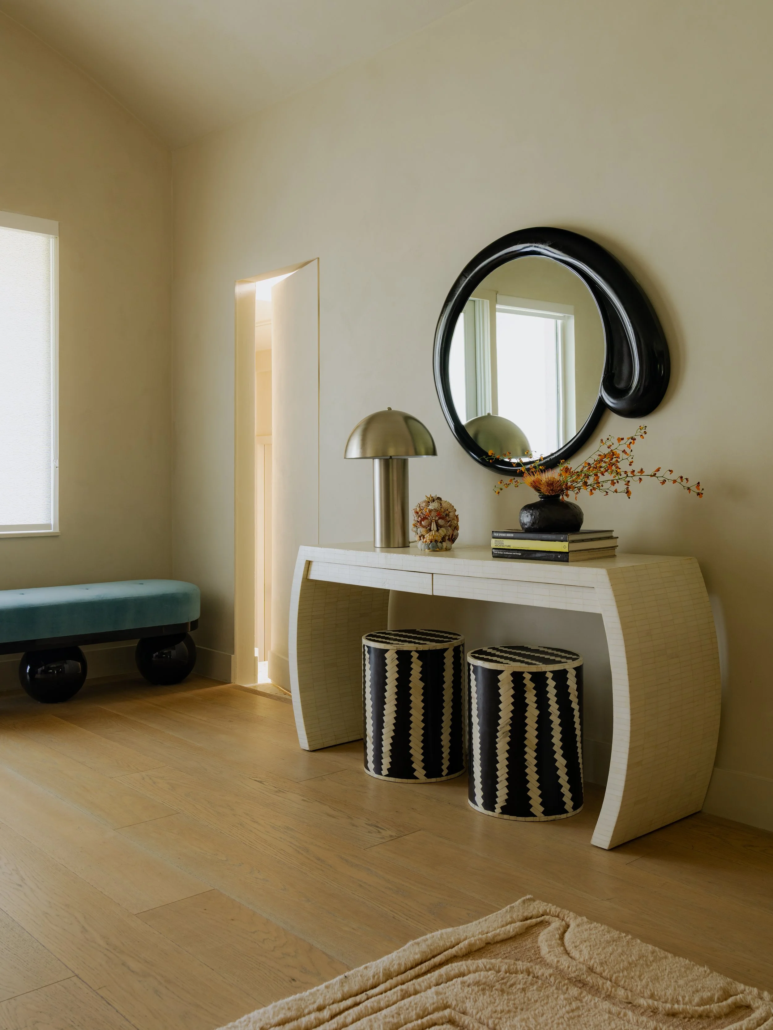 A modern living room corner with a white curved console table, two black and white zigzag patterned stools, a black vase with orange and red flowers, a metallic table lamp, and a black framed oval mirror on a beige wall. There are two windows allowing natural light, a beige textured rug on light wood flooring, and a light-colored doorway leading to another room.