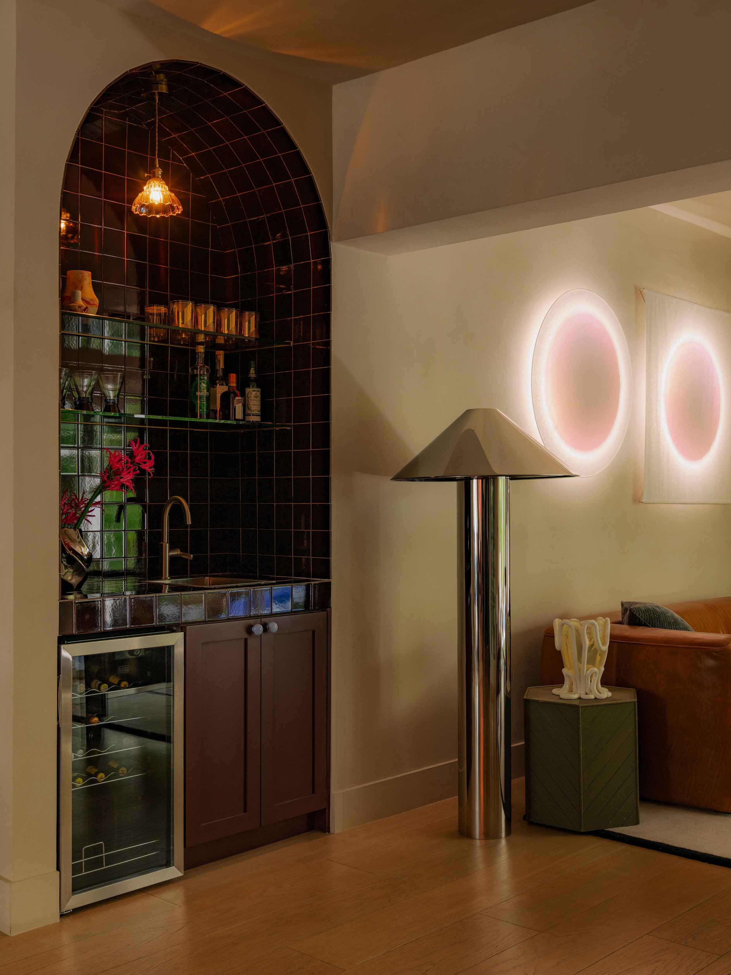 Interior of a room with a wine fridge, a dark wood cabinet, a bar area with bottles and glasses, a modern metallic floor lamp, and a sofa with a side table holding a white sculpture, illuminated by soft wall lighting.