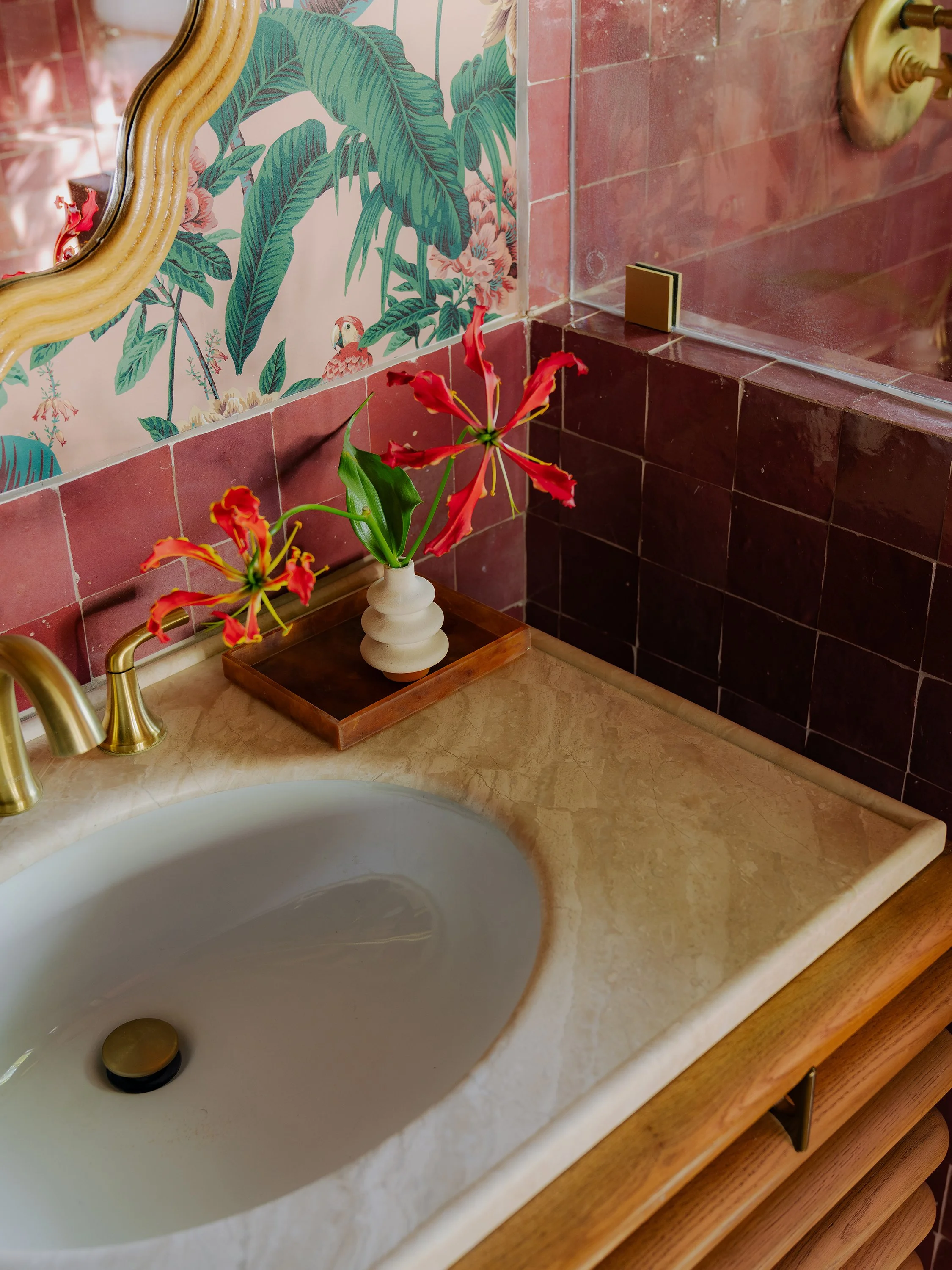 Bathroom sink with a marble countertop, gold faucet, a small vase with red flowers, and a tropical-themed wallpaper with large green leaves and pink flamingos.