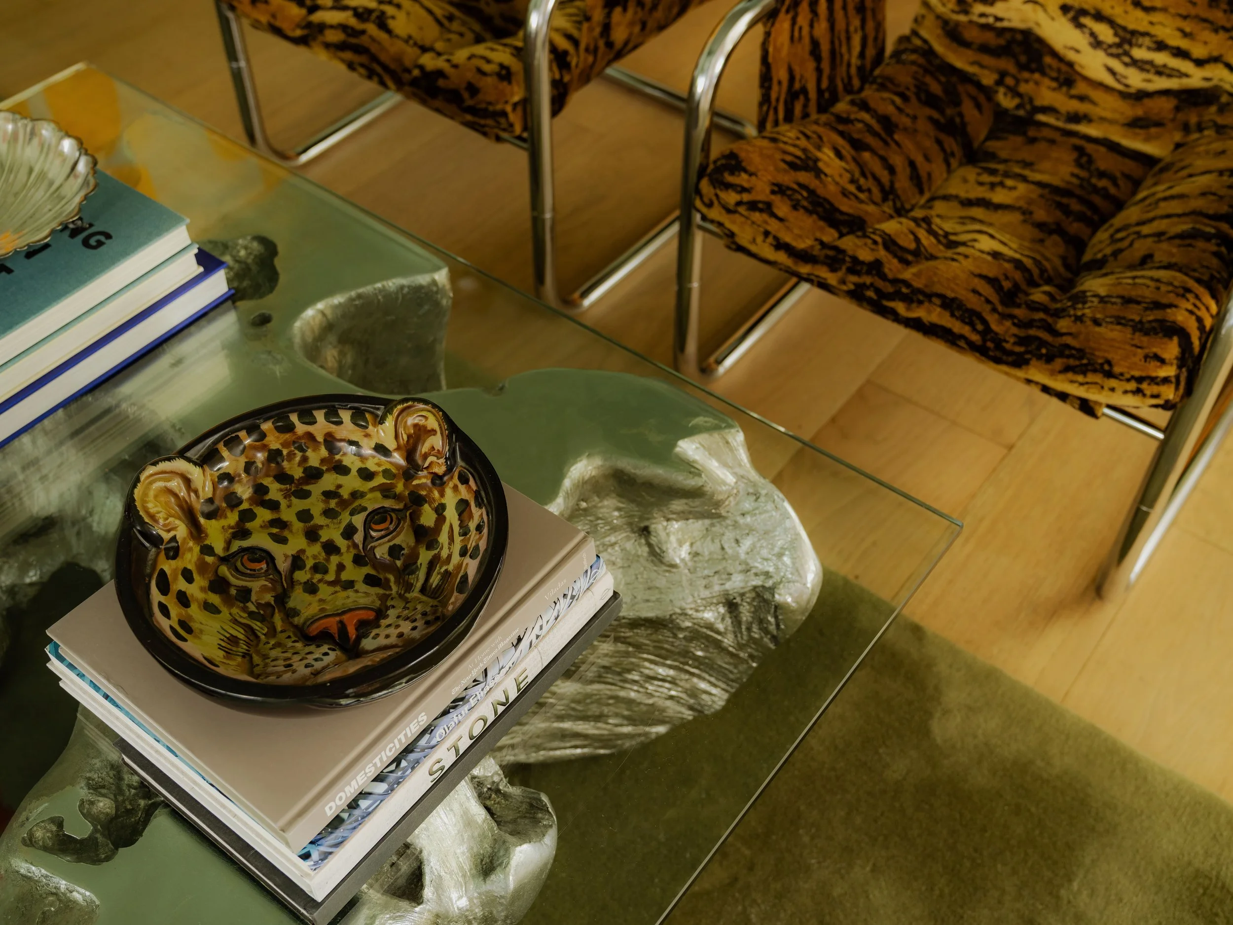 A glass coffee table with a leopard-print ceramic bowl, a stack of books, and decorative rocks on top. In the background, there are two vintage-style chairs with leopard-patterned upholstery and metallic frames.