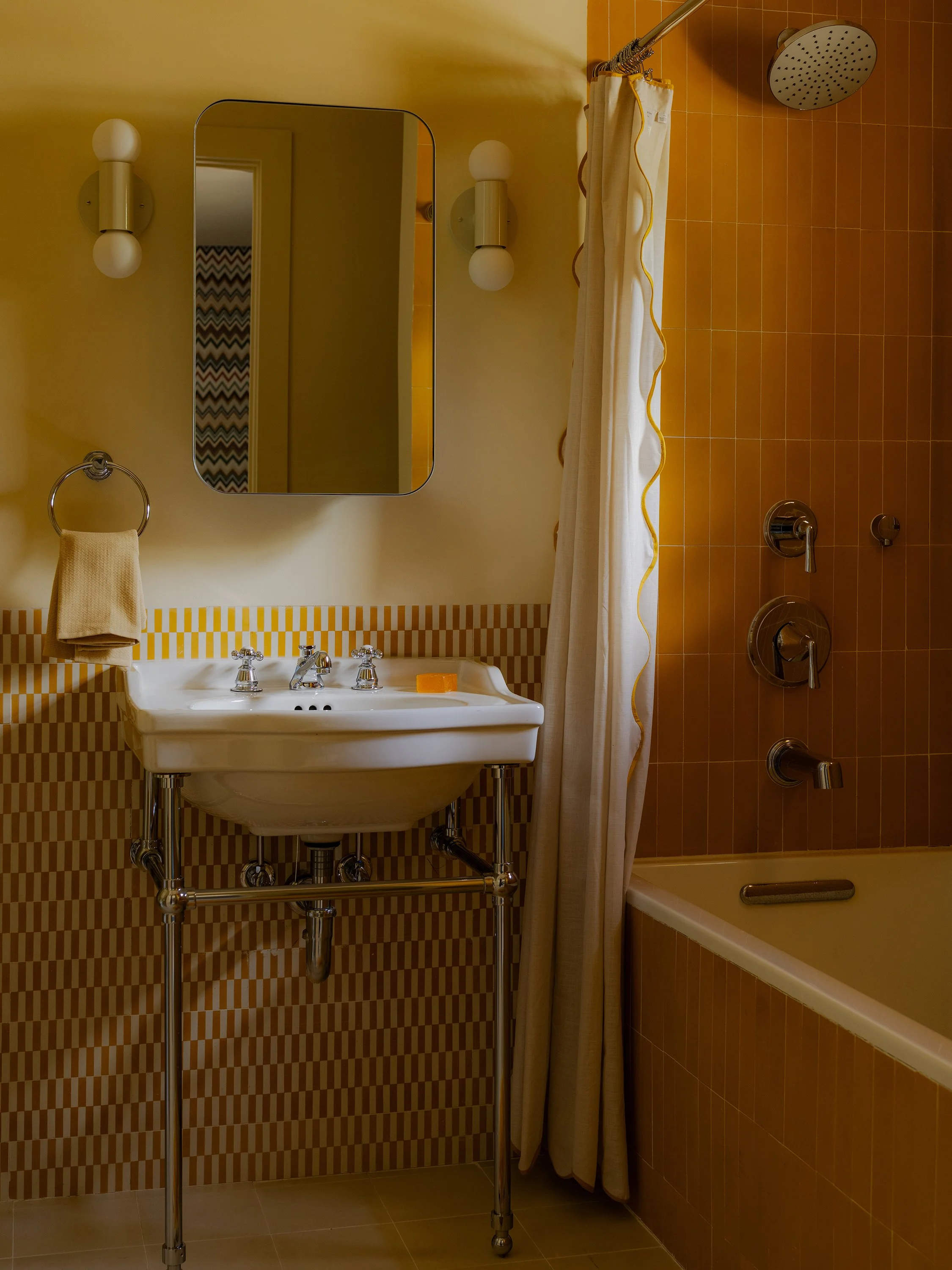 Bathroom with yellow tiles, a white sink with chrome fixtures, a mirror, a wall-mounted light, a towel ring with a towel, a bathtub with a shower curtain, and a showerhead.