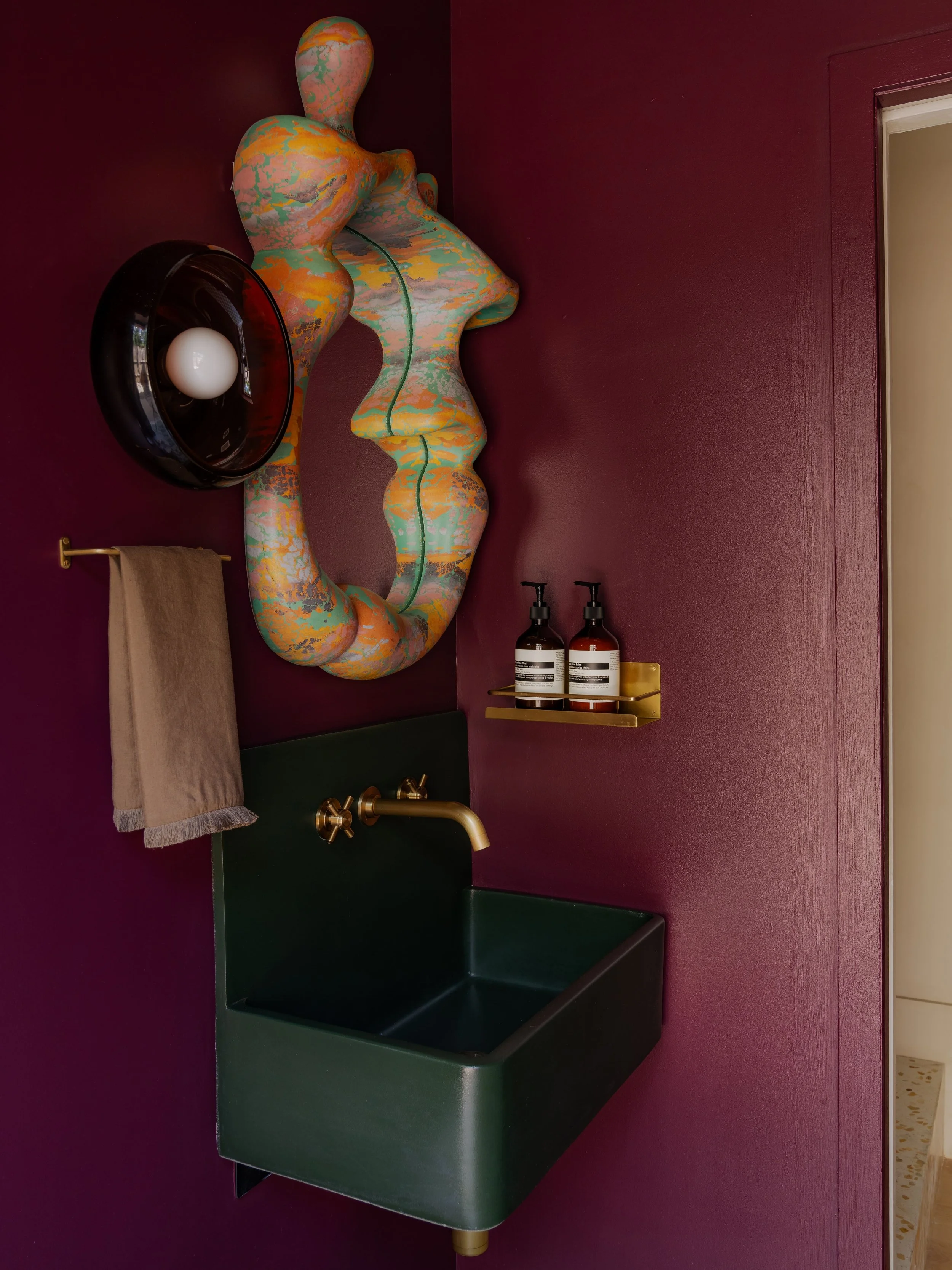 A green bathroom sink with a gold faucet, mounted on a wall painted purple. To the right, two black bottles with soap dispensers and a small yellow item on a gold shelf. To the left, a beige towel hanging on a gold towel bar. Above the sink, a colorful, abstract wall sculpture with organic, flowing shapes in orange, green, and pink hues. Next to the sculpture, a black round light fixture with a white bulb.