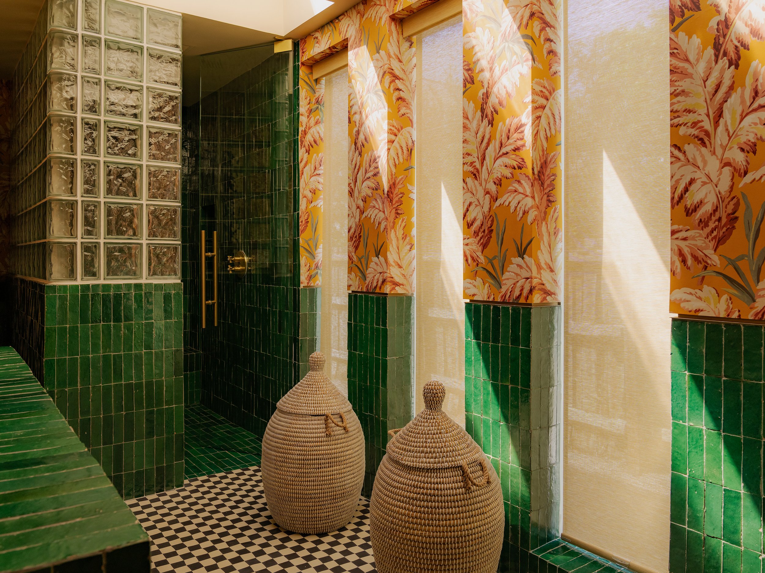 Indoor space with green tiled surfaces, a glass block wall, and patterned curtains with orange and yellow leaves, along with woven baskets on the floor.