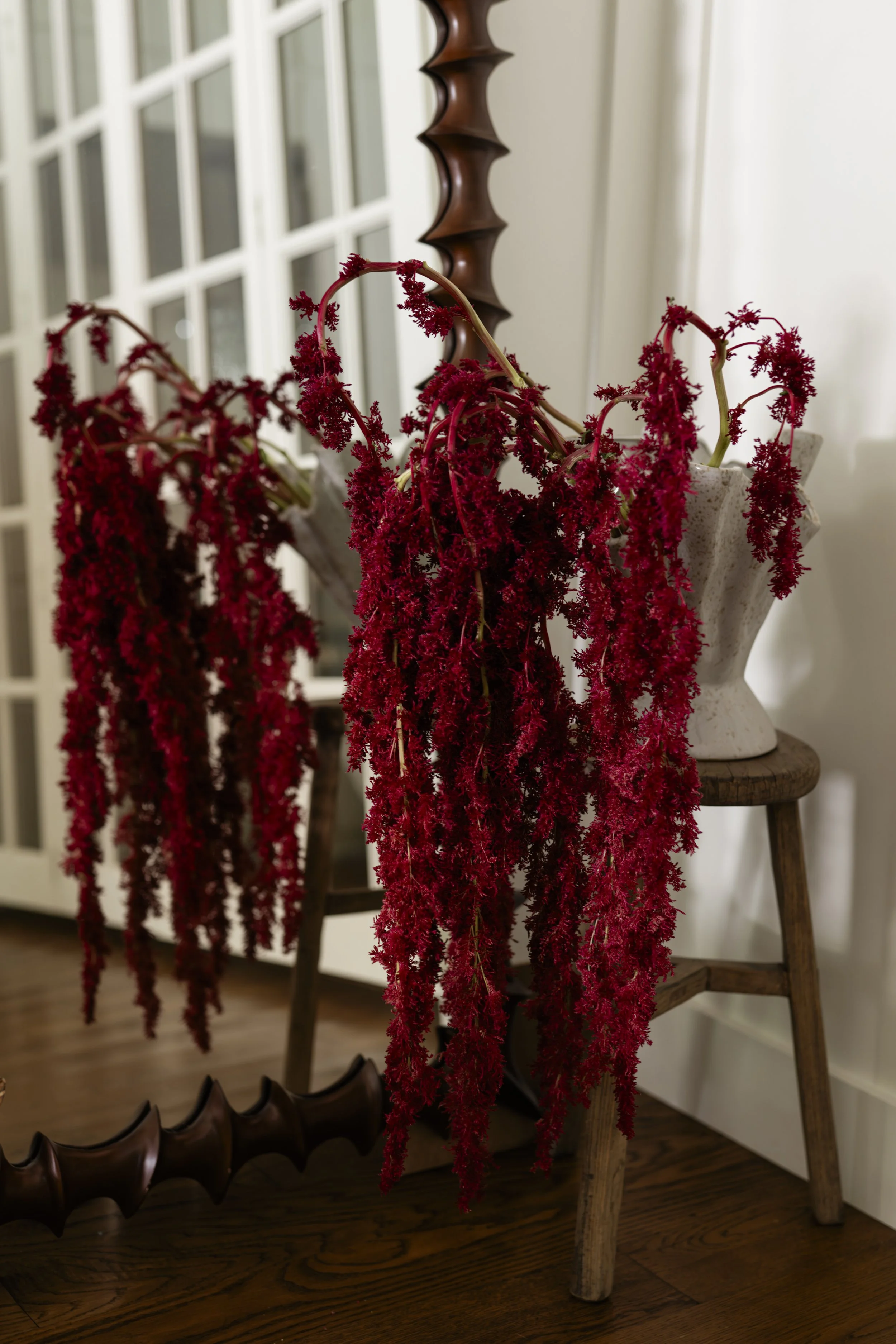 Red hanging amaranth flowers in a white vase on a wooden chair, with brown cowboy boots on the floor nearby, and a wood-paneled floor and white wall background.