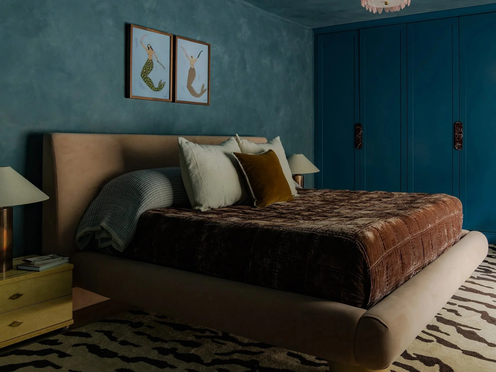 A bedroom with a beige bed, multiple pillows in white and brown, a beige headboard, and a dark brown velvet bedspread. There are two white nightstands with lamps on each side and a yellow drawer nightstand with books and a remote on the right side. The wall behind the bed is teal with two paintings of mermaids, and there is a dark blue wardrobe on the right. A zebra-patterned rug is partly visible on the floor.