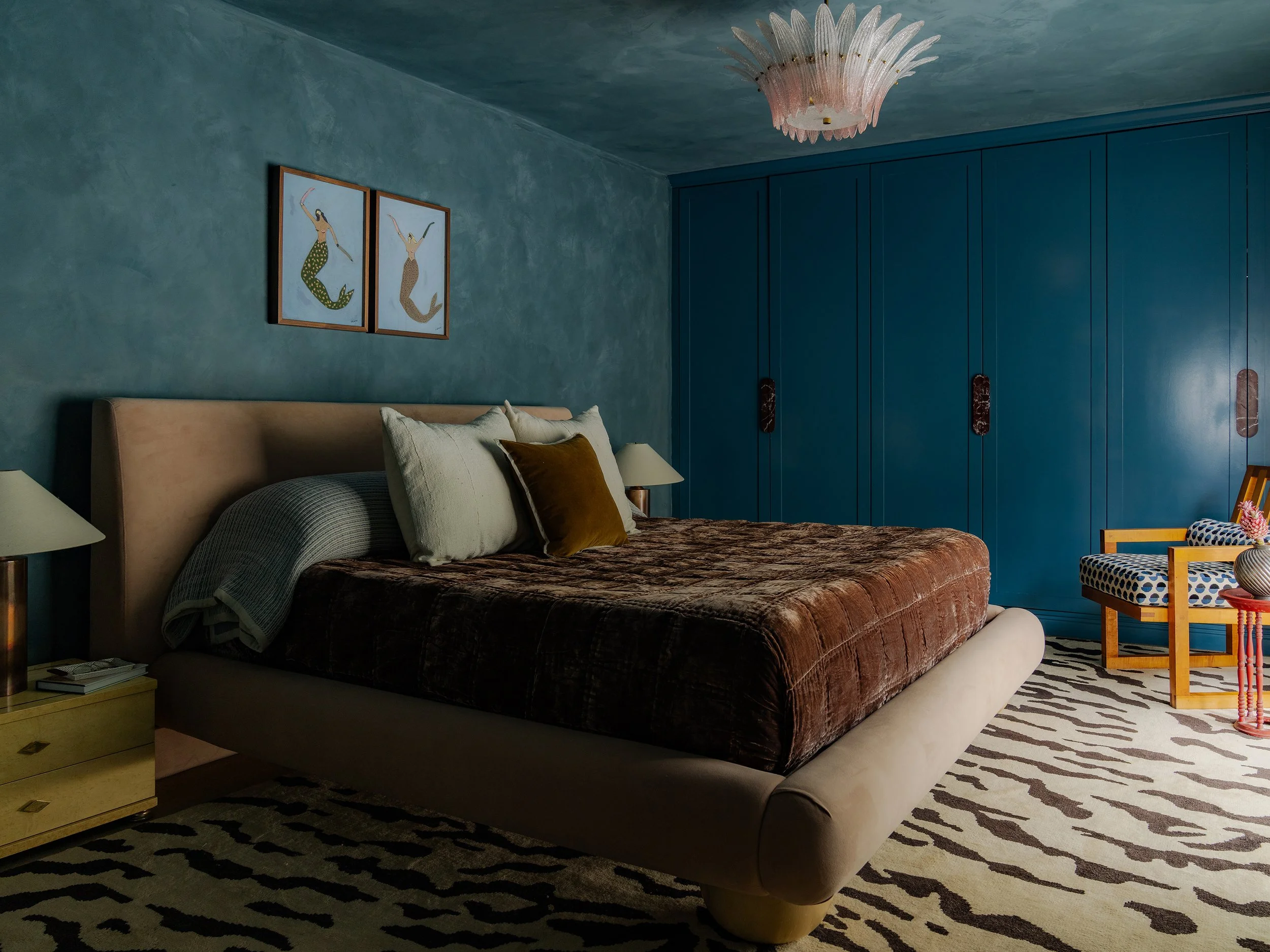 A bedroom with a bed, pillows, and a blanket, a nightstand with books and a lamp, two framed mermaid artwork on the wall, a large blue wardrobe, a patterned rug, and a chair with a polka dot cushion.
