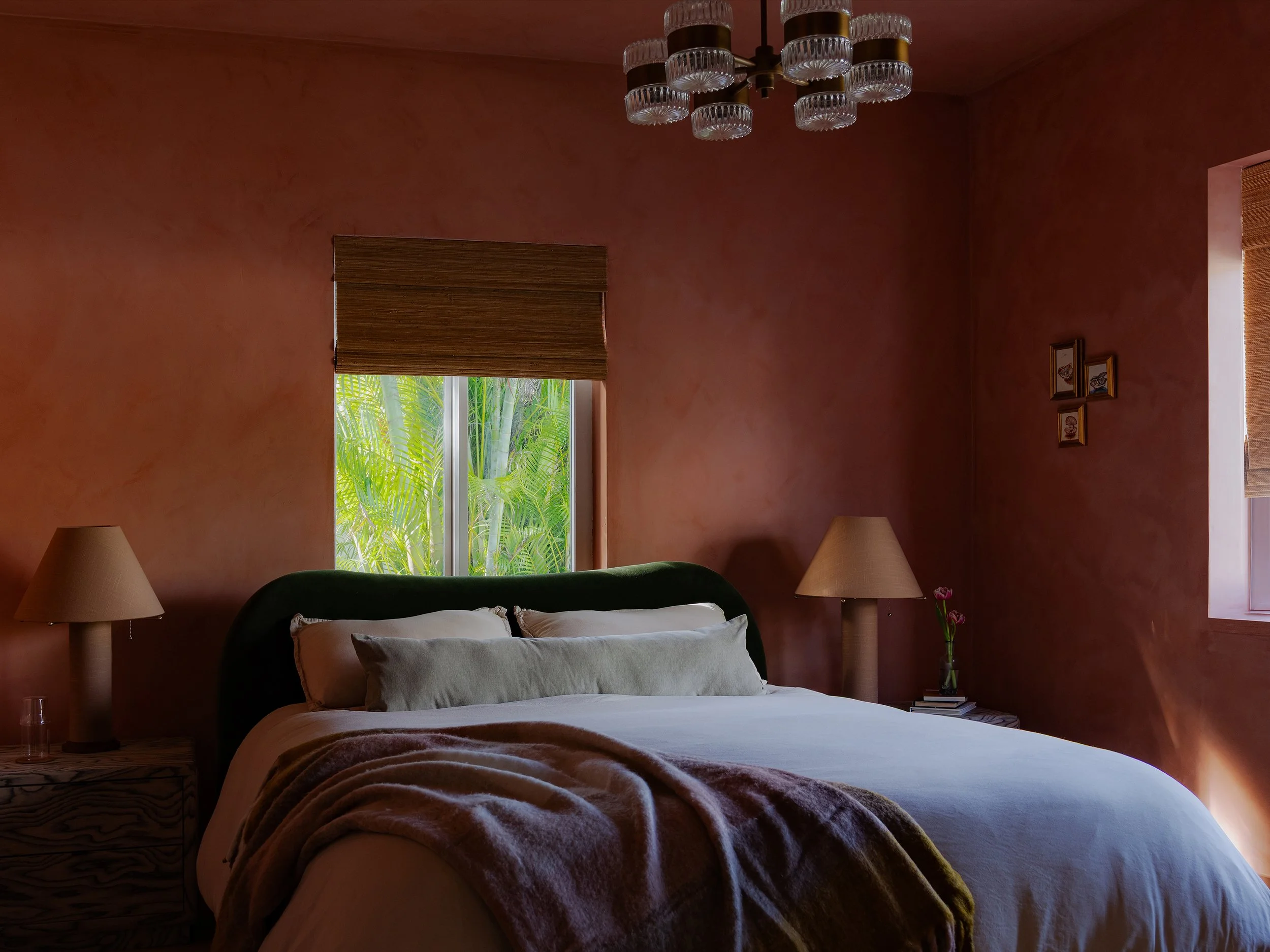 A cozy bedroom with a large bed covered in white and brown blankets, two nightstands with lamps, a window with a bamboo shade, and a view of lush green foliage outside.