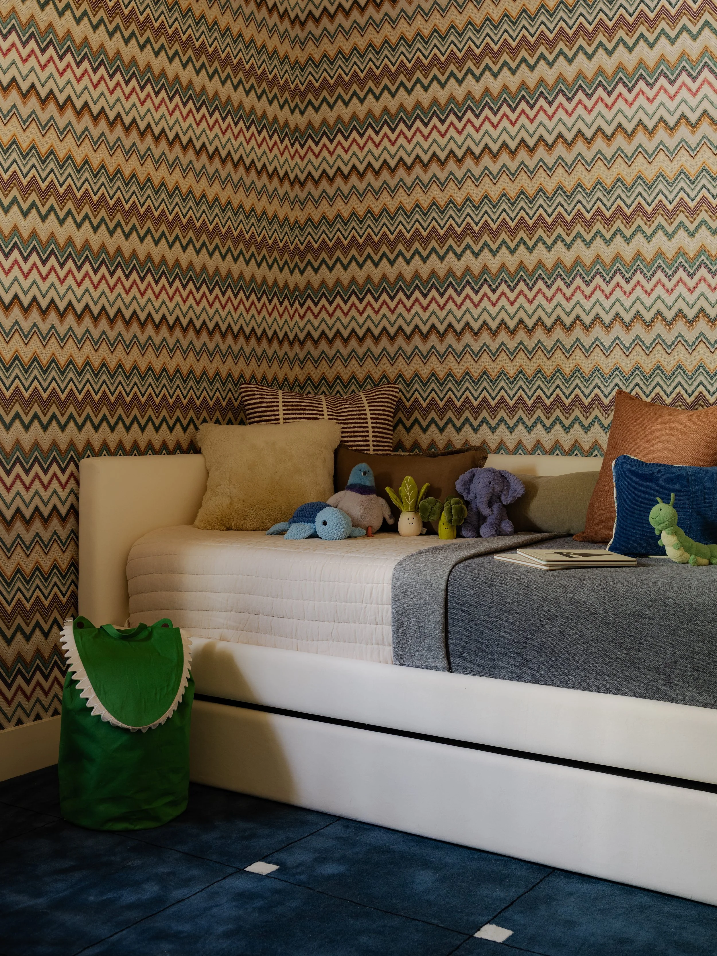 A white bed with various stuffed animals and pillows, a green bag shaped like a crocodile on the floor, and a textured, zigzag-patterned wall in the background.