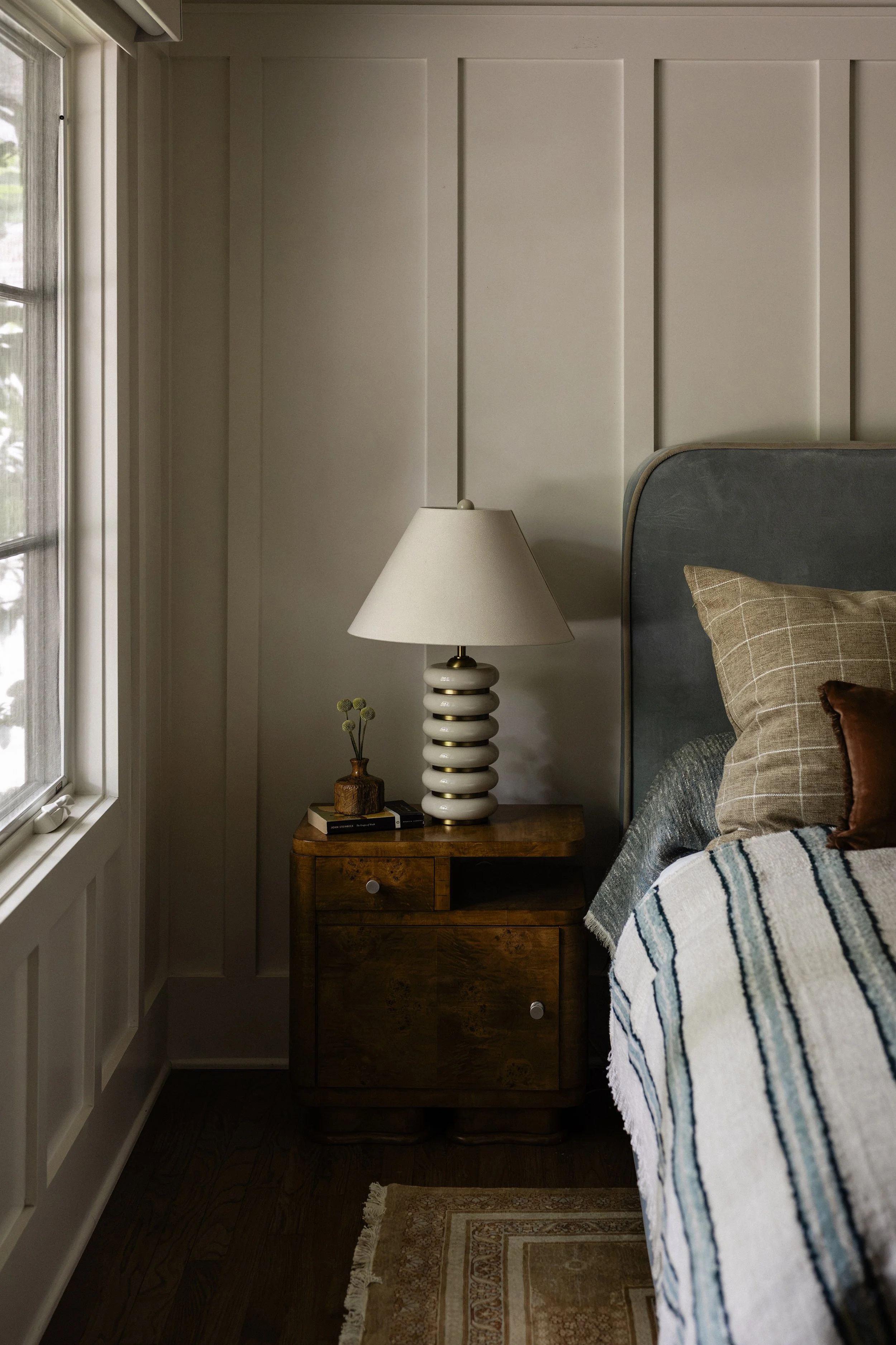 A bedroom corner featuring a wooden nightstand with a cream-colored, wavy ceramic lamp and a small vase with dried flowers. Part of the bed with a checked pillow and blanket is visible. The wall has vertical paneling, and there is a window to the left with a sheer curtain, allowing natural light to enter.