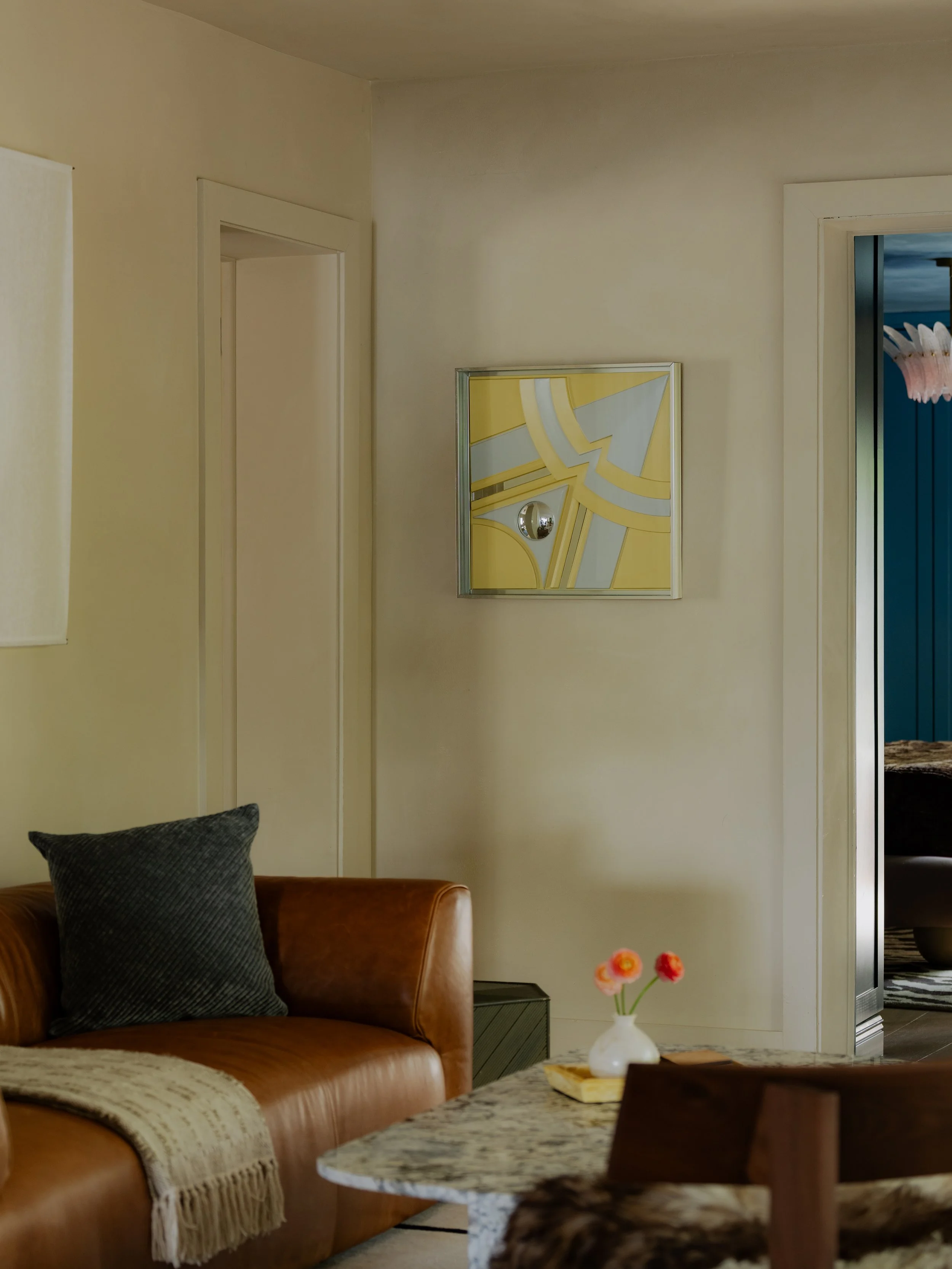Living room interior with a brown leather sofa, a dark pillow, a marble tabletop with a white vase holding pink and orange flowers, beige walls, a framed abstract yellow and gray artwork, and an open doorway revealing a blue room with a chandelier.