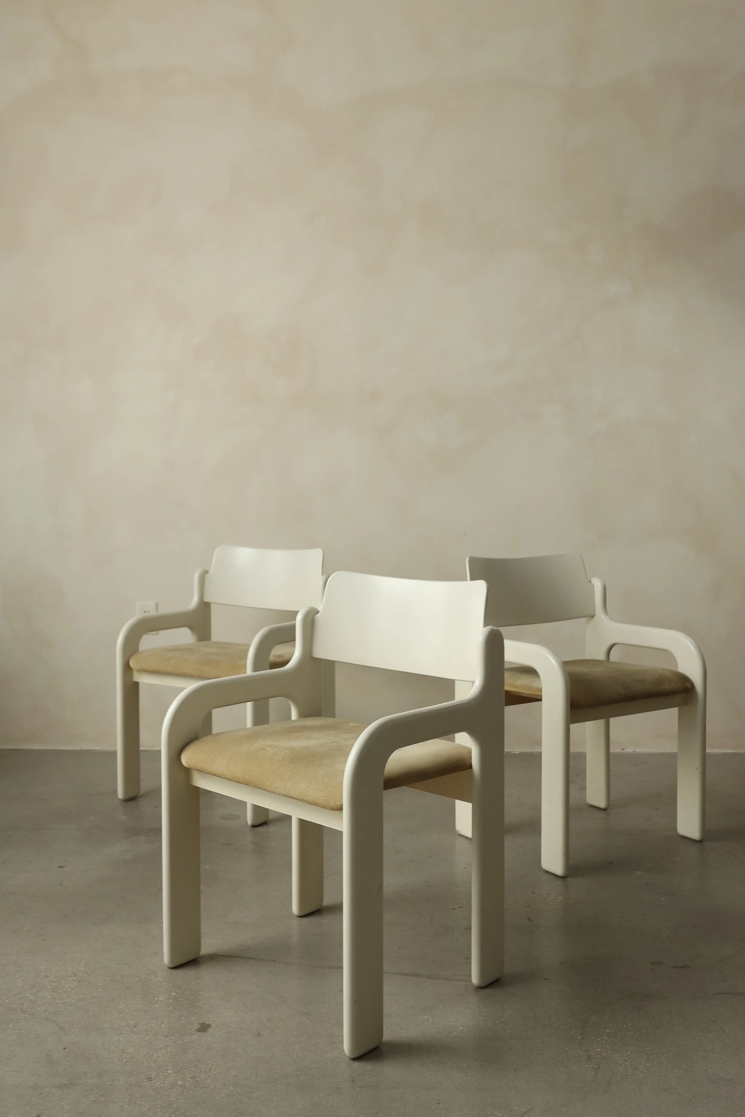 Four white chairs with beige cushions arranged in a row against a plain beige wall.