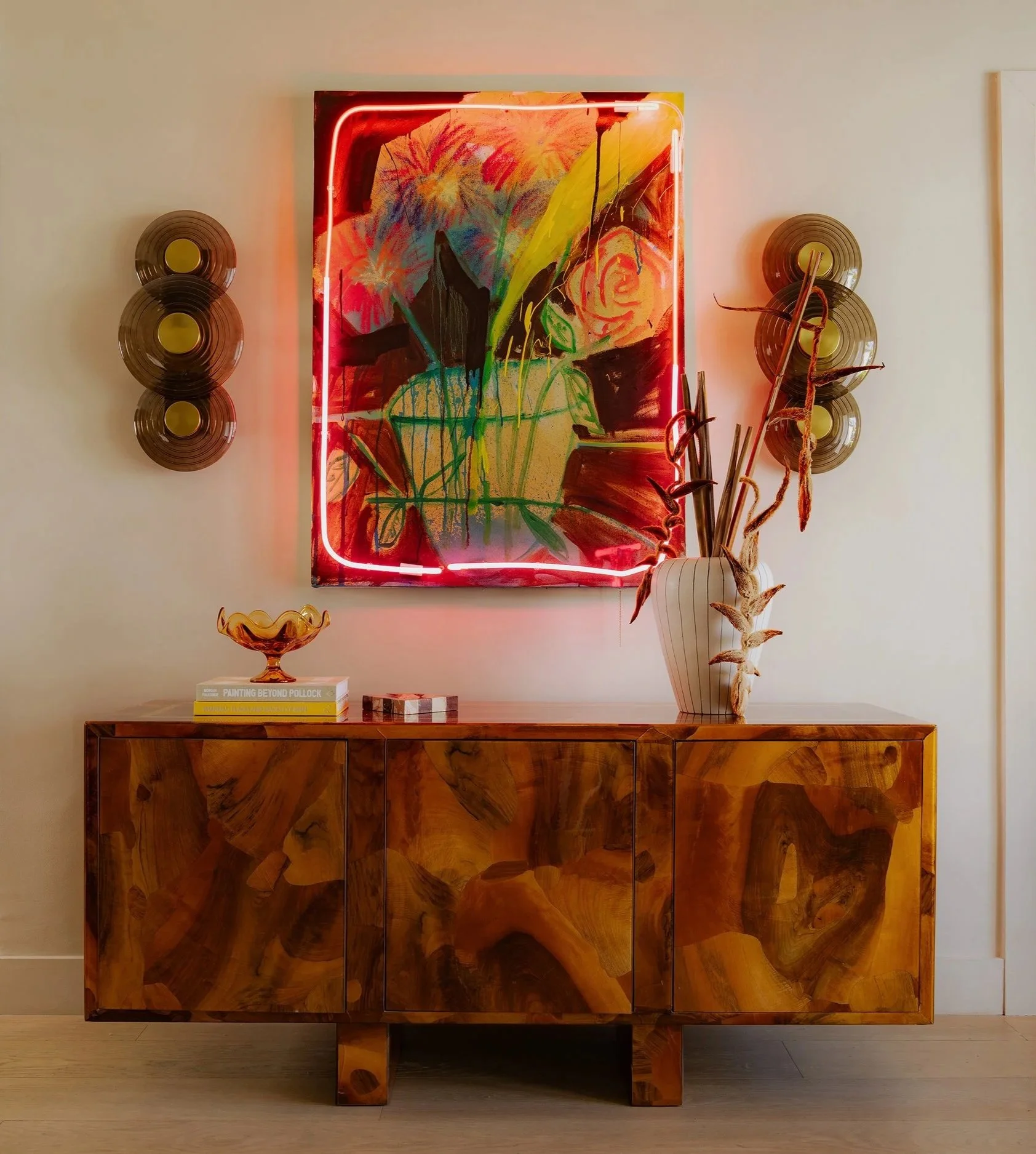 A wooden cabinet with a glossy finish under a colorful floral painting with a neon light frame, flanked by two wall-mounted circular decor pieces, and a decorative bowl with books and a sculpture on top.