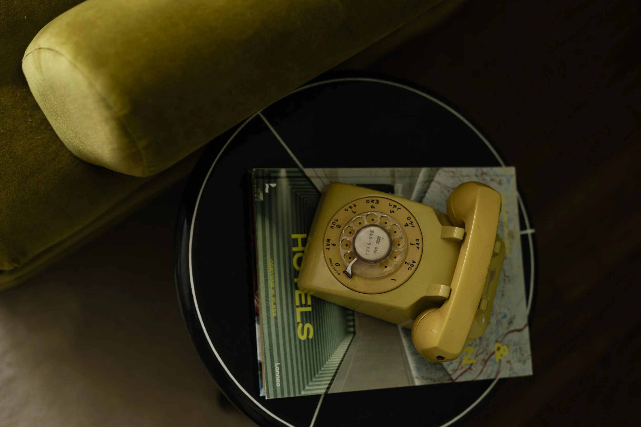 A yellow rotary dial phone placed on top of a rolled-up map near a book and a magazine, on a round black table with white trim.