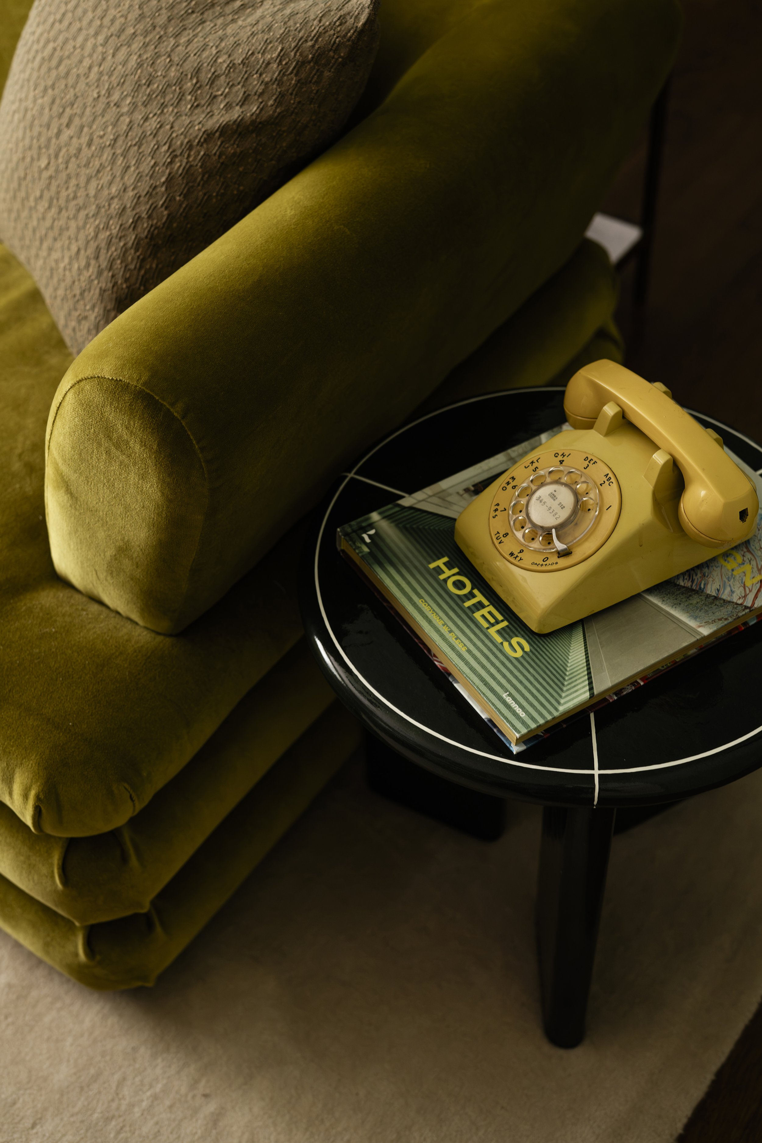 A yellow rotary telephone resting on top of a magazine on a black circular table, next to a green upholstered armchair.
