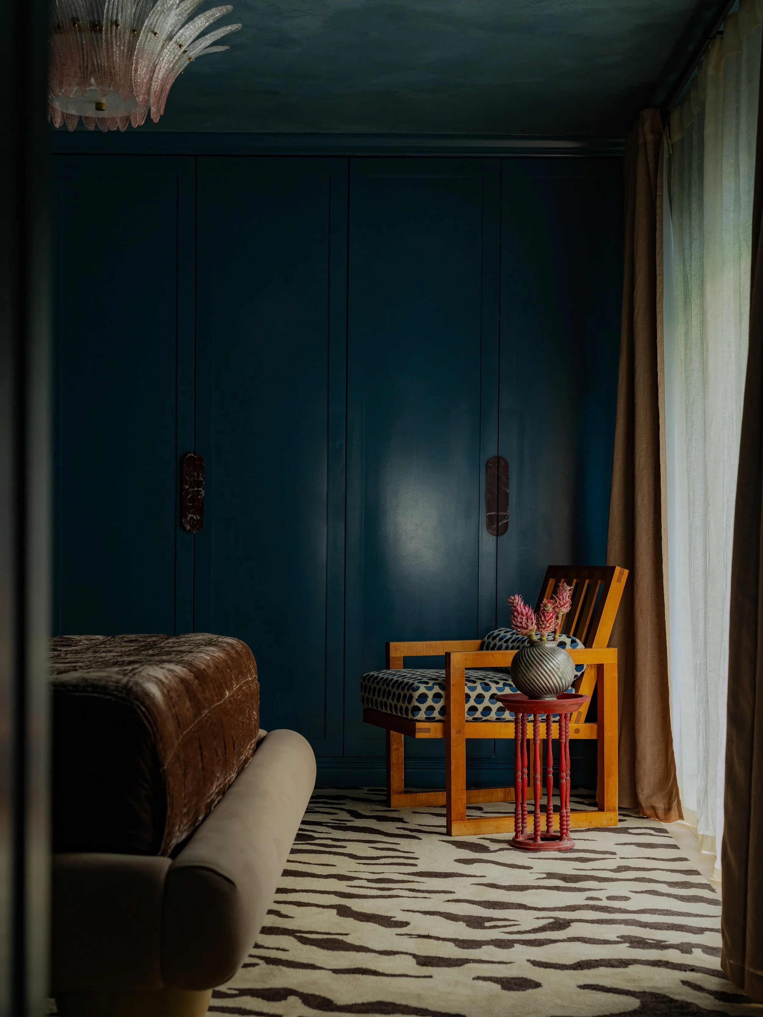 Living room corner with blue built-in cabinets, a brown velvet sofa, a patterned armchair, a vase with pink flowers on a red side table, wooden furniture, a zebra-patterned rug, and sheer curtains by a window.
