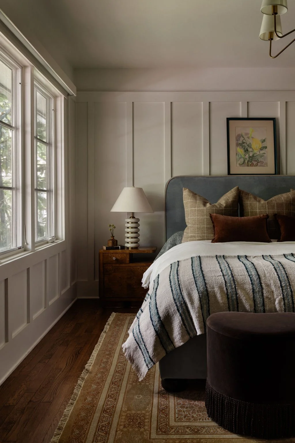 A cozy bedroom with a bed featuring a gray headboard, brown and checkered pillows, and a striped blanket. There's a wooden nightstand with a distinctive white lamp, a framed botanical artwork on the wall, a window with white trim letting in natural light, and part of a dark-colored chair in the foreground. The floor has a patterned rug and wooden flooring.