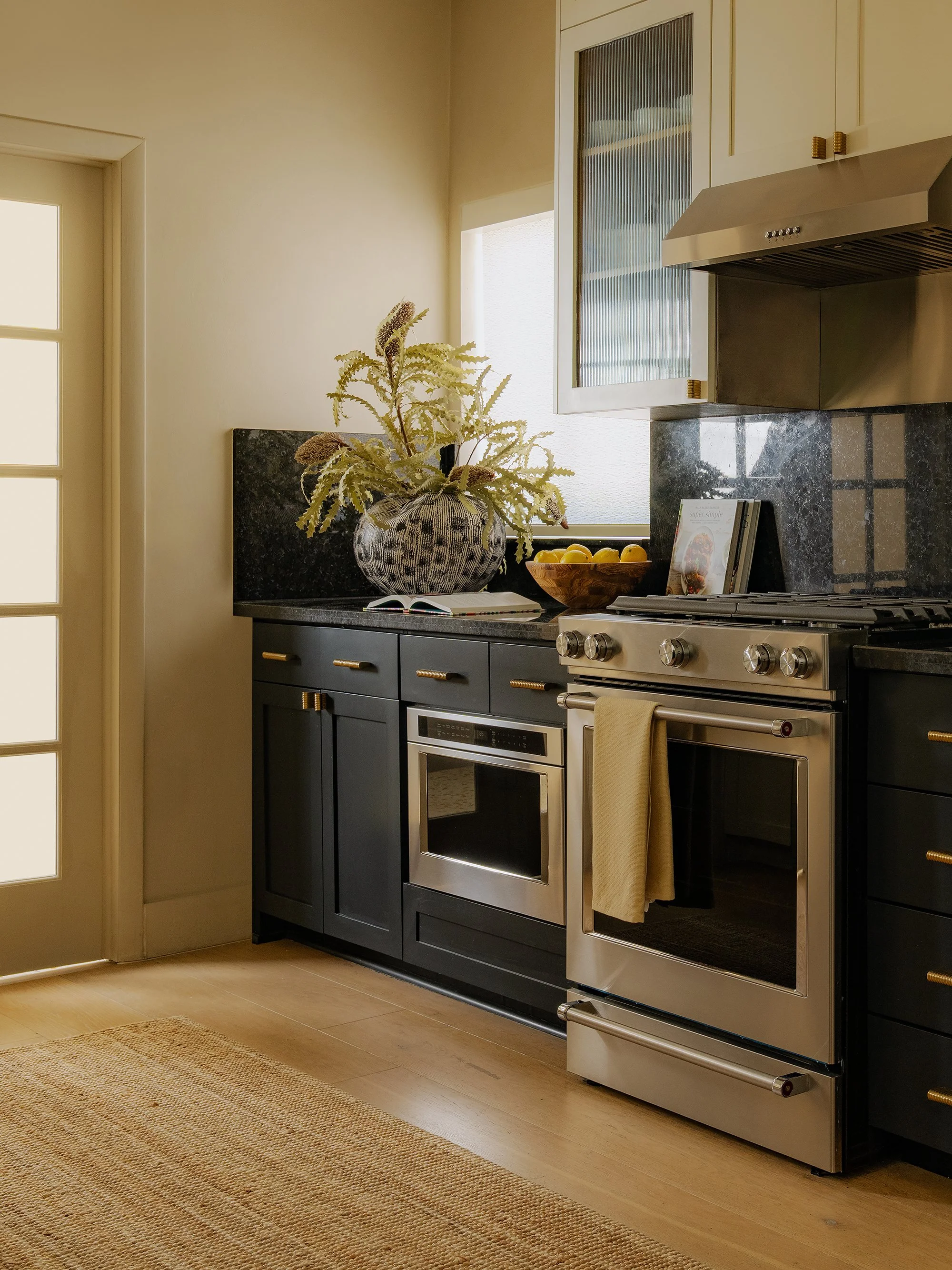 A kitchen with black cabinets, a stainless steel oven and stove, beige walls, and a window above the stove. There is a large decorative vase with a plant, a bowl of lemons, and a cookbook on the counter. A beige rug is on the wooden floor.