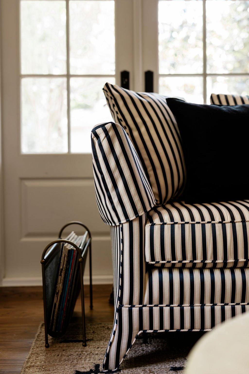 Close-up of a black and white striped armchair with cushion, next to a standing magazine rack near a window in a well-lit room.