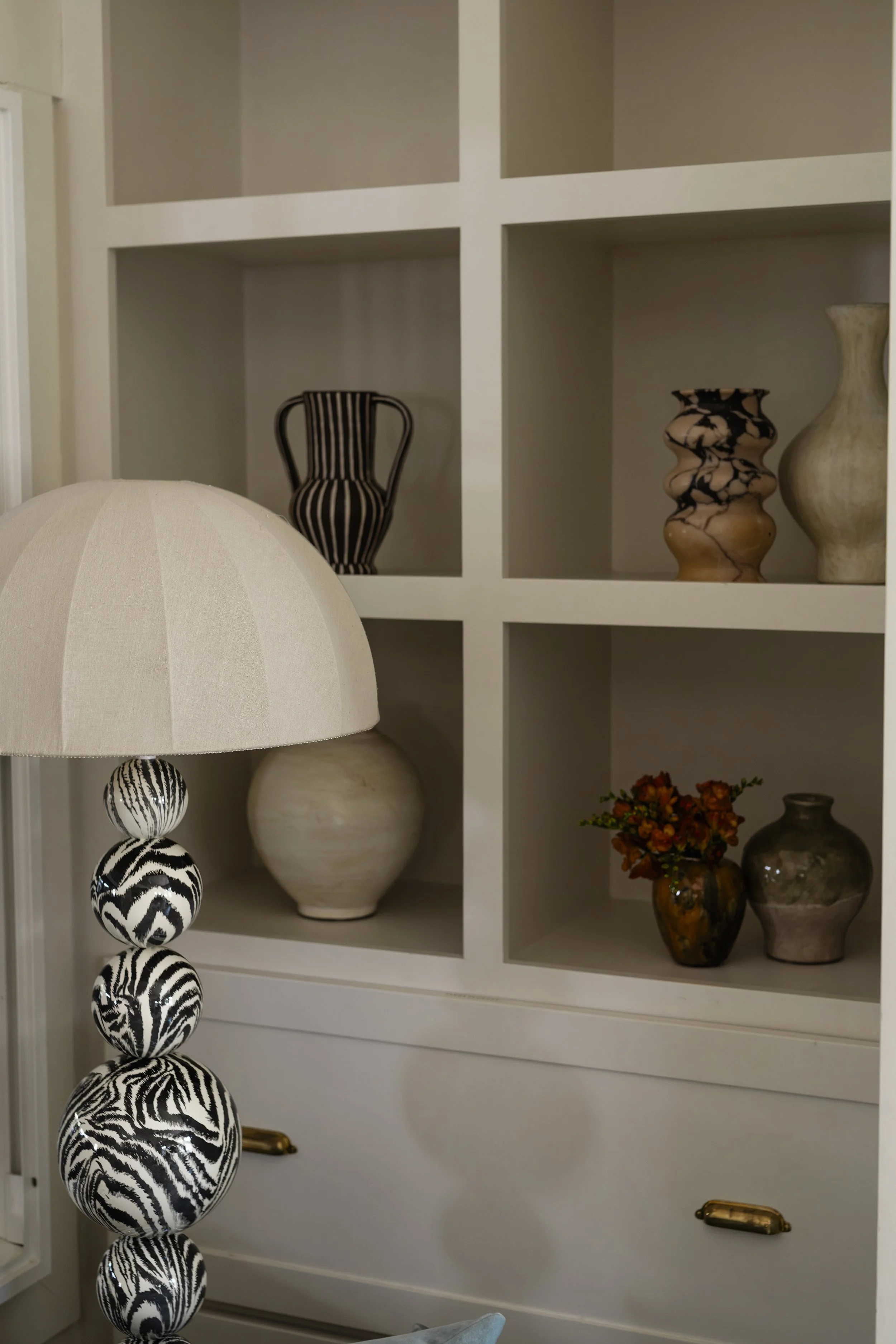 Decorative vases on the shelves of a white built-in cabinet, with a portion of a black and white beaded hanging decoration and a white table lamp visible in the foreground.
