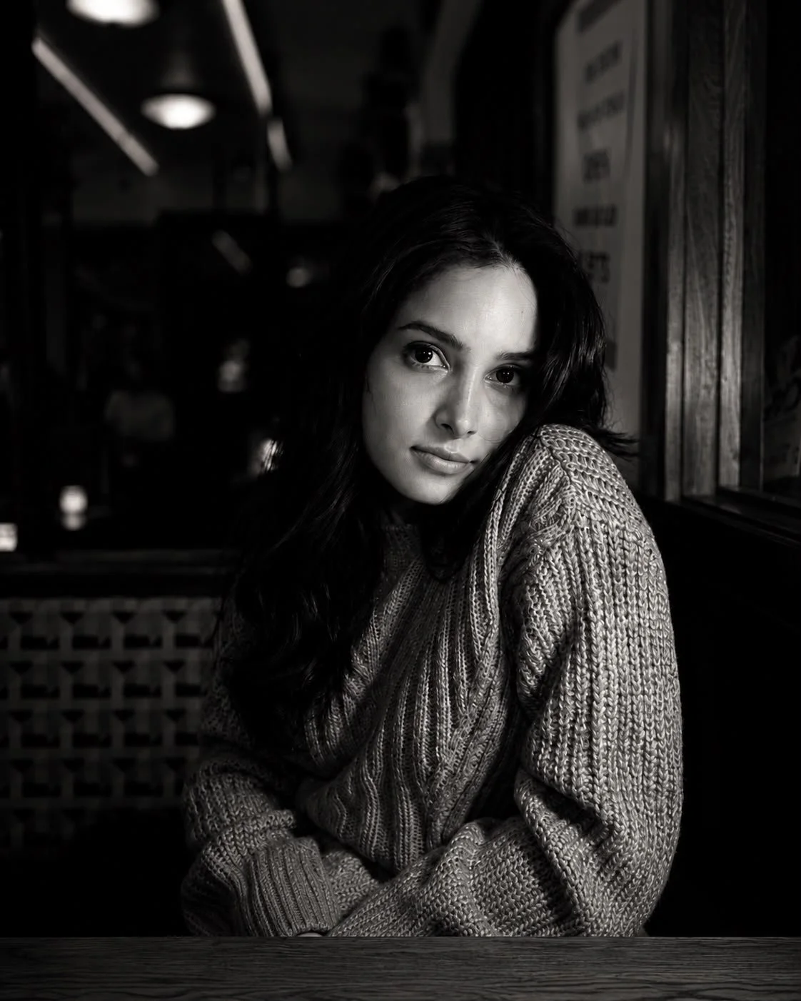 A black and white portrait of a woman sitting indoors near a window, wearing a textured sweater, with long dark hair and a contemplative expression.