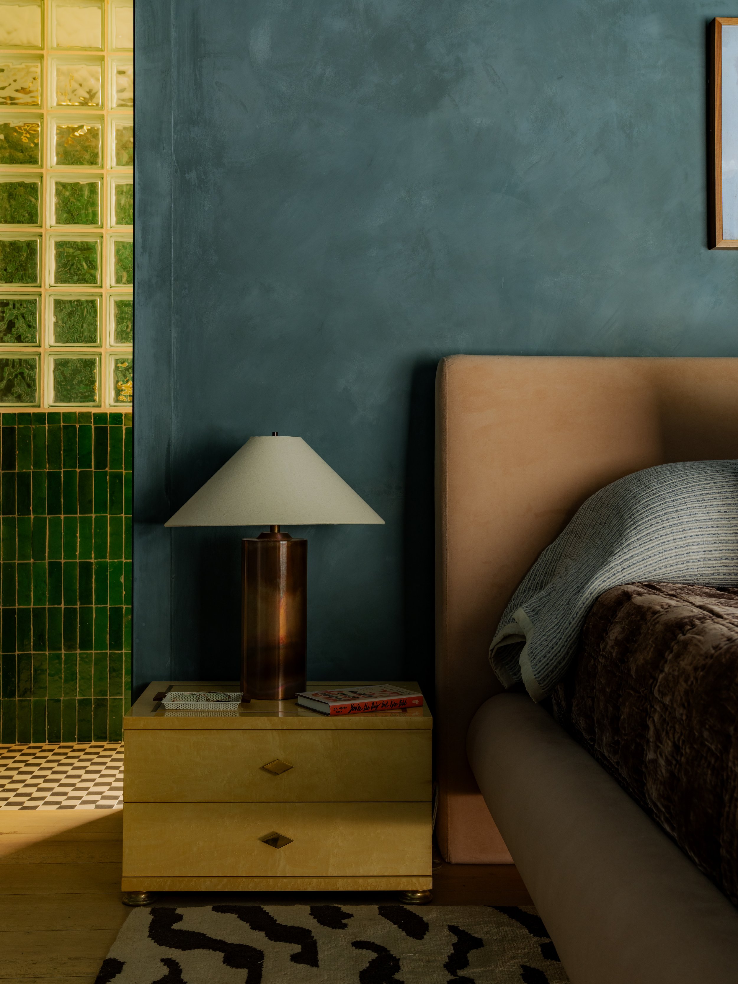 A bedroom corner with a beige bed, a yellow nightstand holding a lampshade and books, a blue wall, and a glass block window with yellow-glazed glass blocks.