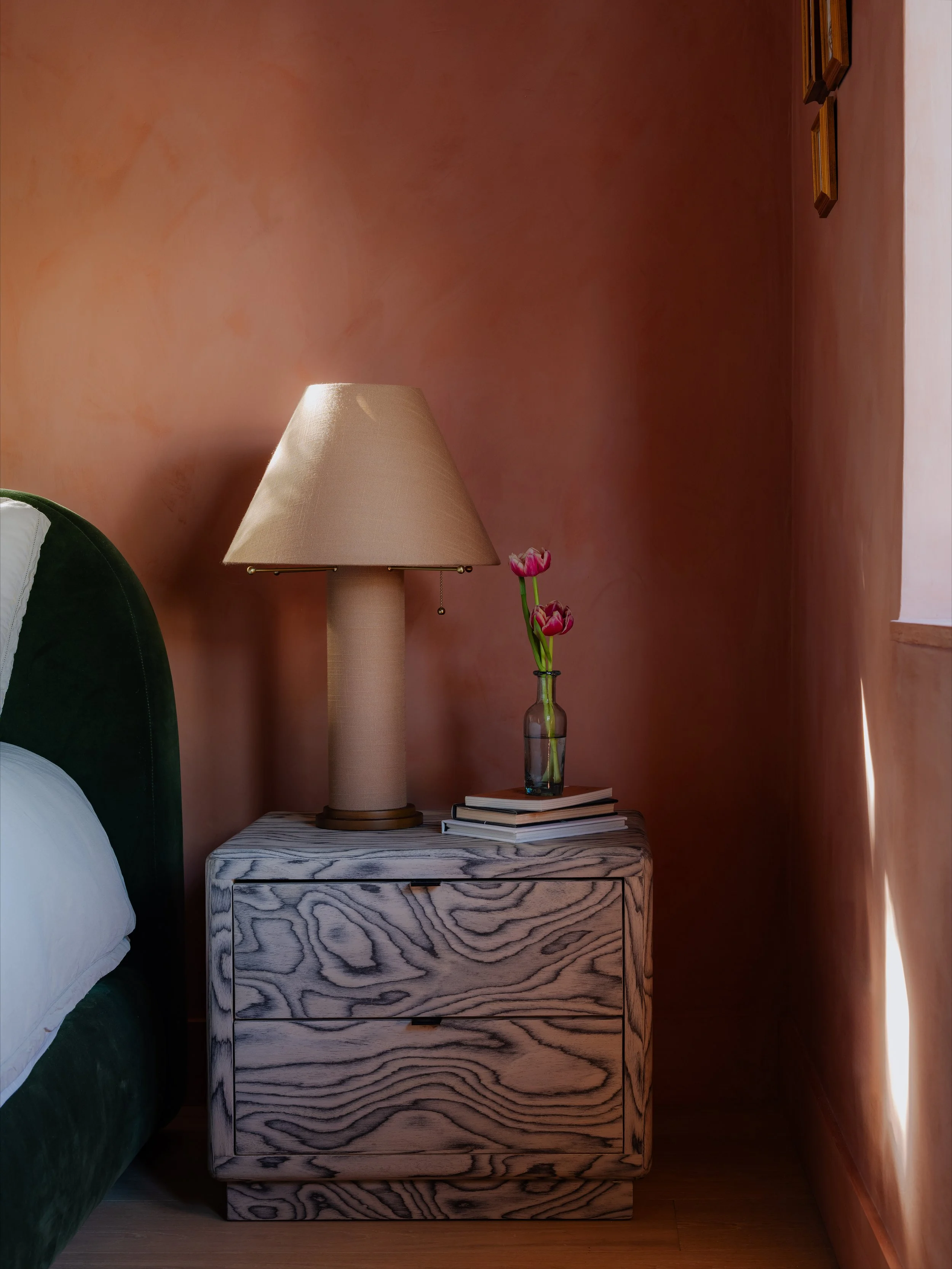 A bedside table with shadowed wood grain pattern, holding a tan table lamp, a glass vase with pink tulips, and a stack of books, outside a bedroom with pinkish walls and framed artwork.