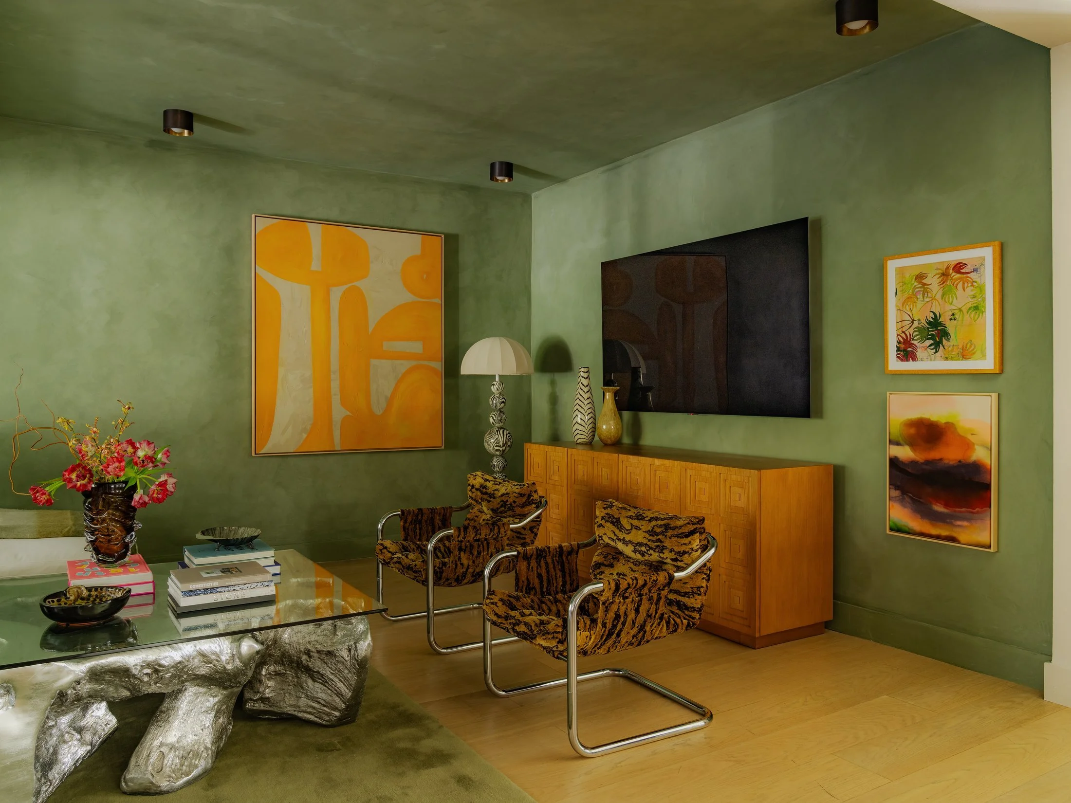 Living room interior with green walls and ceiling, decorated with colorful abstract paintings, a floral arrangement on a glass coffee table, gold-colored chairs with animal print upholstery, and a wooden sideboard with vases and lamps.