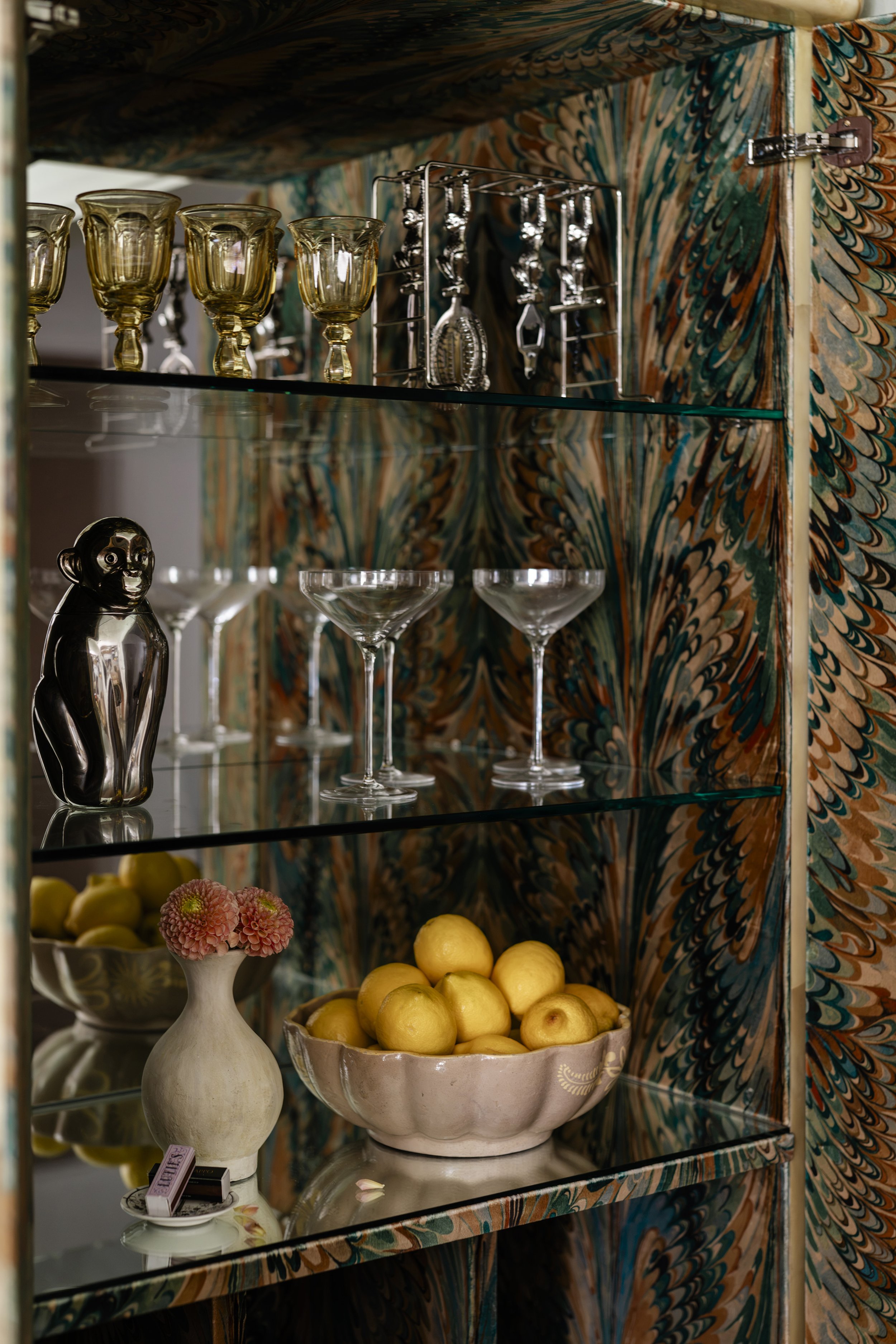 A glass-front cabinet with three shelves. The top shelf contains four yellow wine glasses and a metal jewelry stand. The middle shelf contains decorative items including a ceramic dog sculpture, a small flower arrangement, and a bowl of lemons. The background features a colorful, patterned wallpaper.