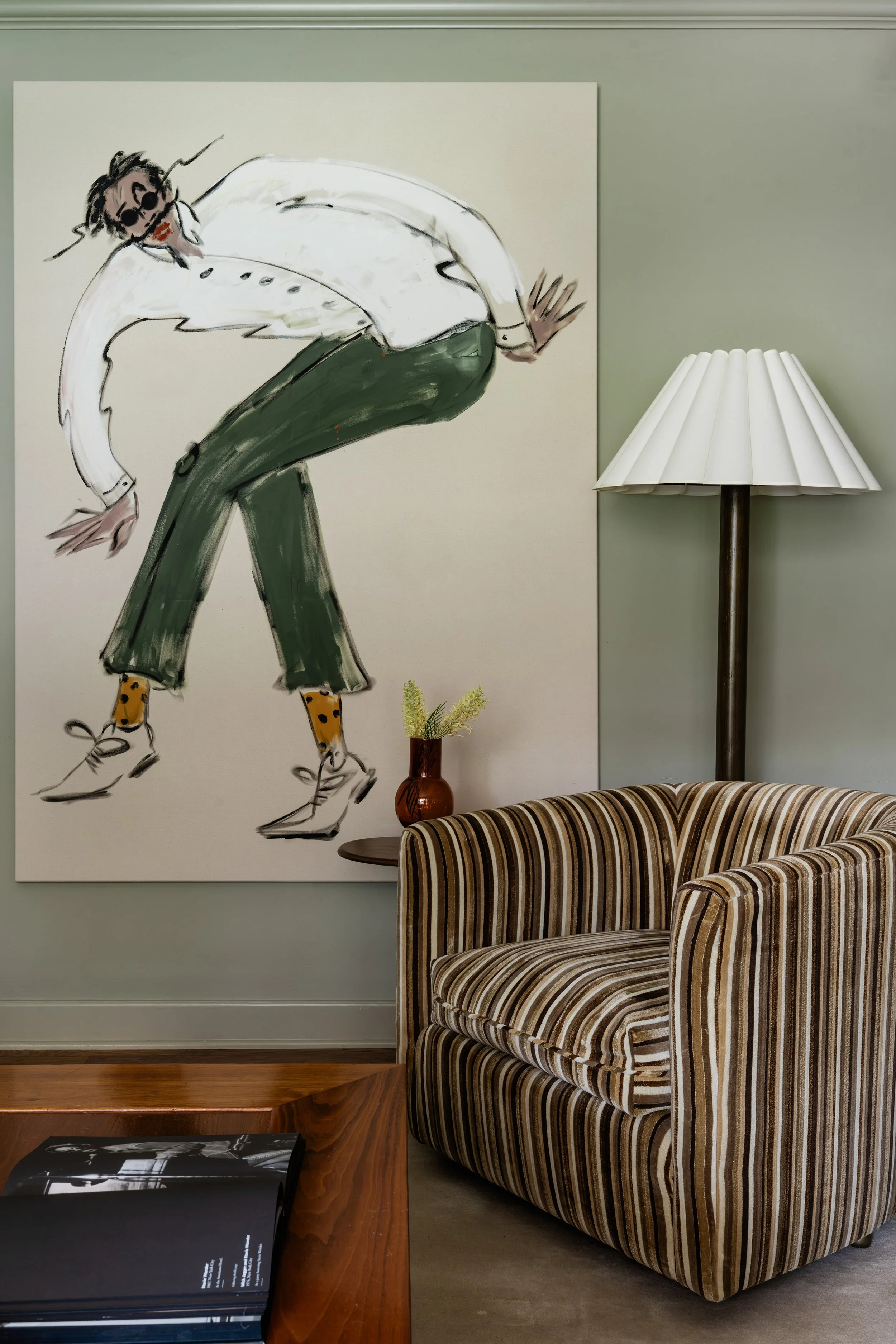 A large painting of a person with sunglasses, white shirt, green pants, and yellow socks standing and leaning forward, displayed on a green wall. A striped armchair is in the foreground, with a wooden side table holding a brown vase with greenery, and a tall floor lamp with a white pleated shade to the right of the painting.