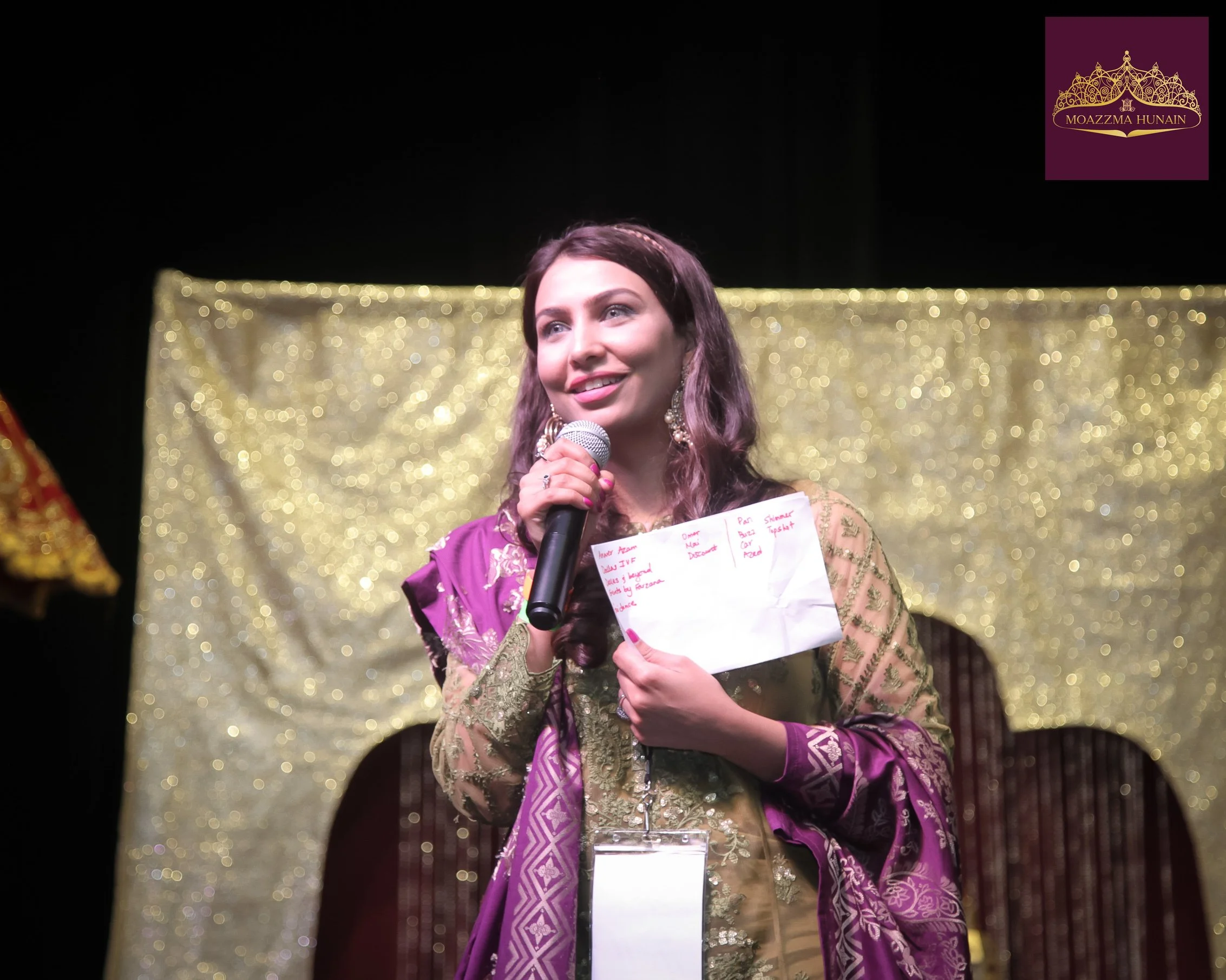 A woman in traditional clothing holding a microphone and a piece of paper, standing in front of a gold, glittery backdrop, with a crown logo that reads 'Moazzma Hunain' in the top right corner.