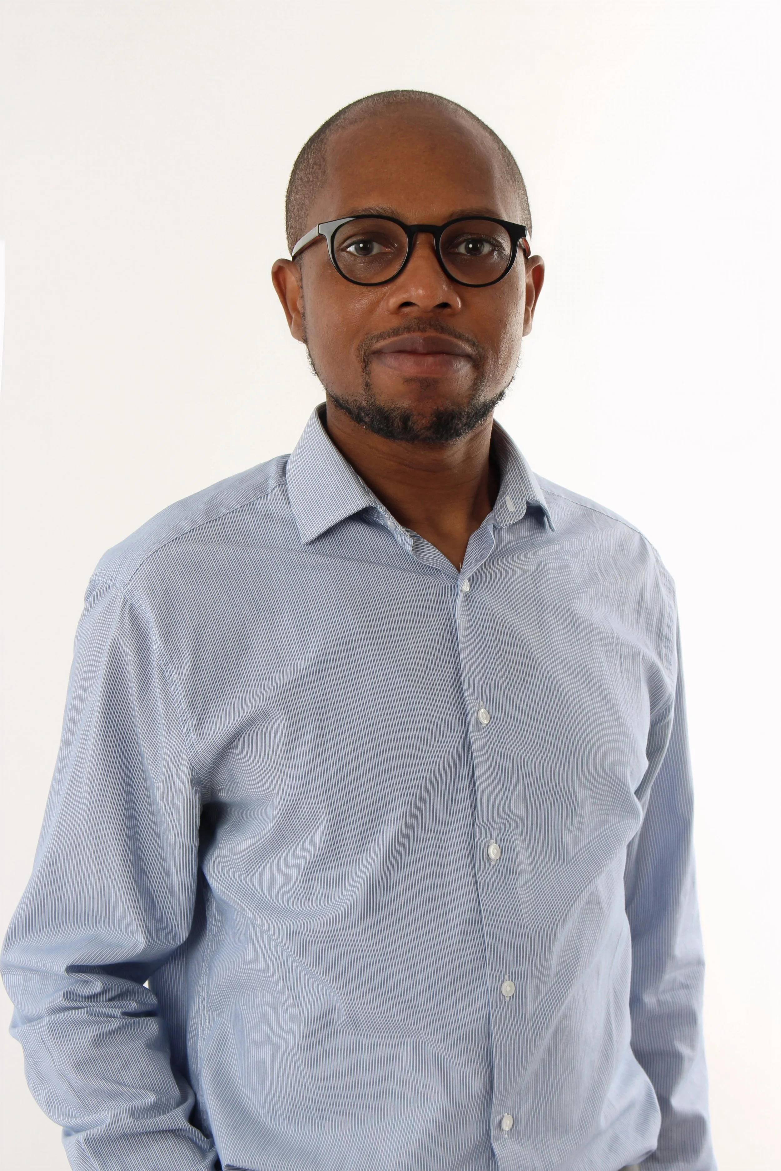 A man with dark skin, glasses, a mustache and beard, wearing a light blue striped button-up shirt, standing against a plain white background.