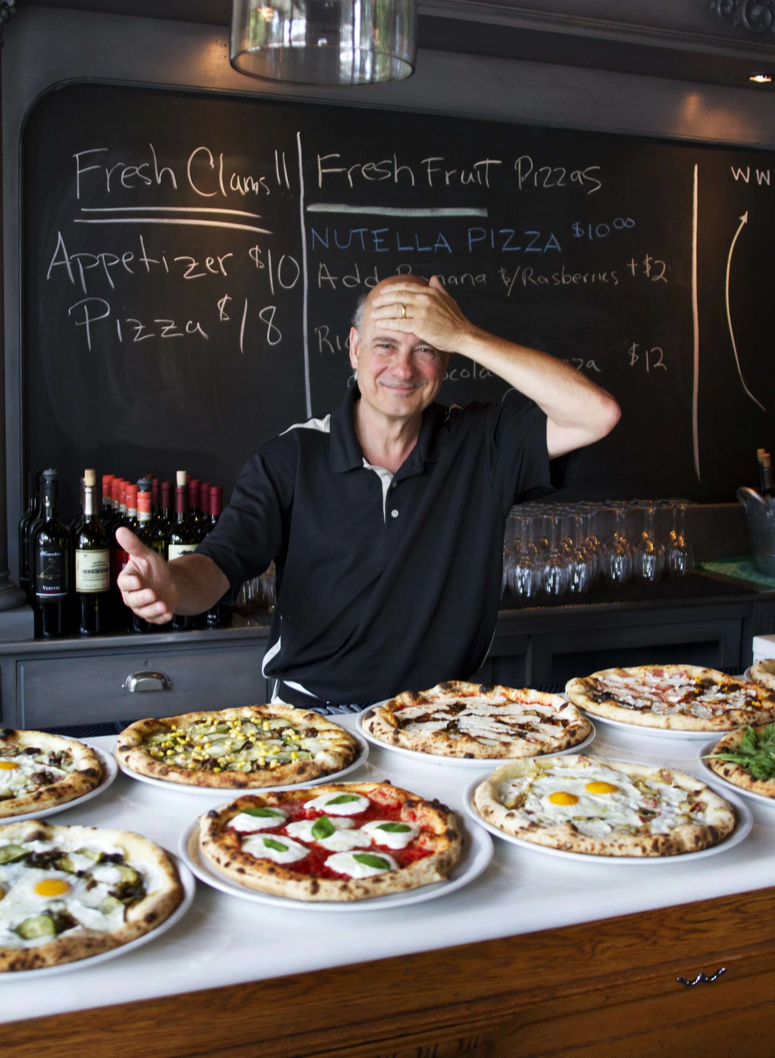 A smiling man with gray hair is standing behind a table of assorted pizzas, in a restaurant or pizzeria. The man is wearing a black polo shirt and appears to be happy. The background includes a chalkboard menu with writings about fresh clams, fruit pizzas, and wine bottles.