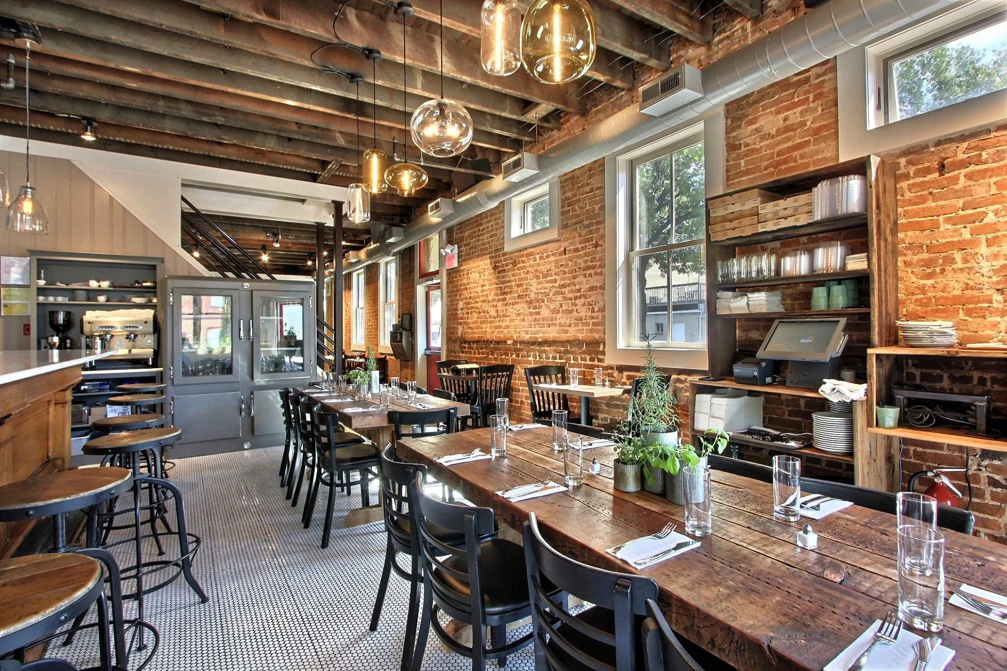 Interior of a rustic restaurant with exposed brick walls, large windows, and wooden ceiling beams. There are tables set with glasses, napkins, and utensils, and a shelf with plates and glassware. Modern hanging lights illuminate the space.