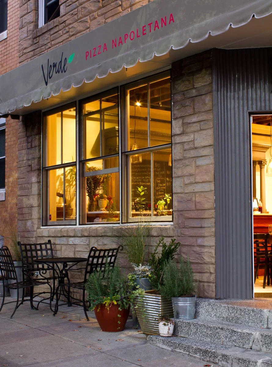 Exterior of a restaurant called Verde with a green awning that has pink and black lettering, reading 'Pizza Napoletana.' The restaurant has large windows showing an warmly lit interior, with potted plants visible inside. Outside, there are black metal chairs and tables, along with various potted plants on the sidewalk, next to steps leading into the restaurant.