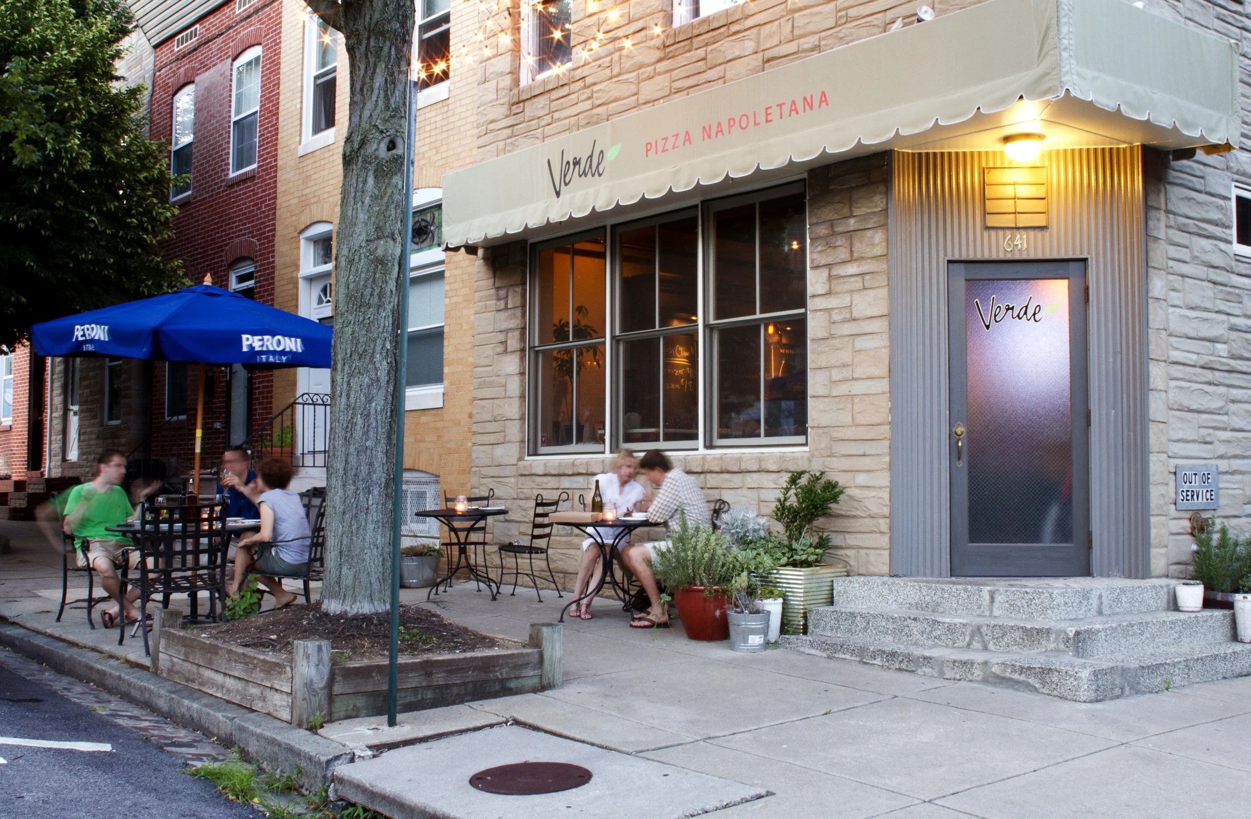 Outdoor seating area in front of a restaurant with a beige awning labeled 'Verde Pizza Napoletana' and a window. There are several tables and chairs with people dining, and a blue umbrella labeled 'Peroni Italy'. A tree blocks part of the scene, and the restaurant has a stone exterior. The sidewalk has a small lawn and plants, and there is a sign indicating the business is out of service.