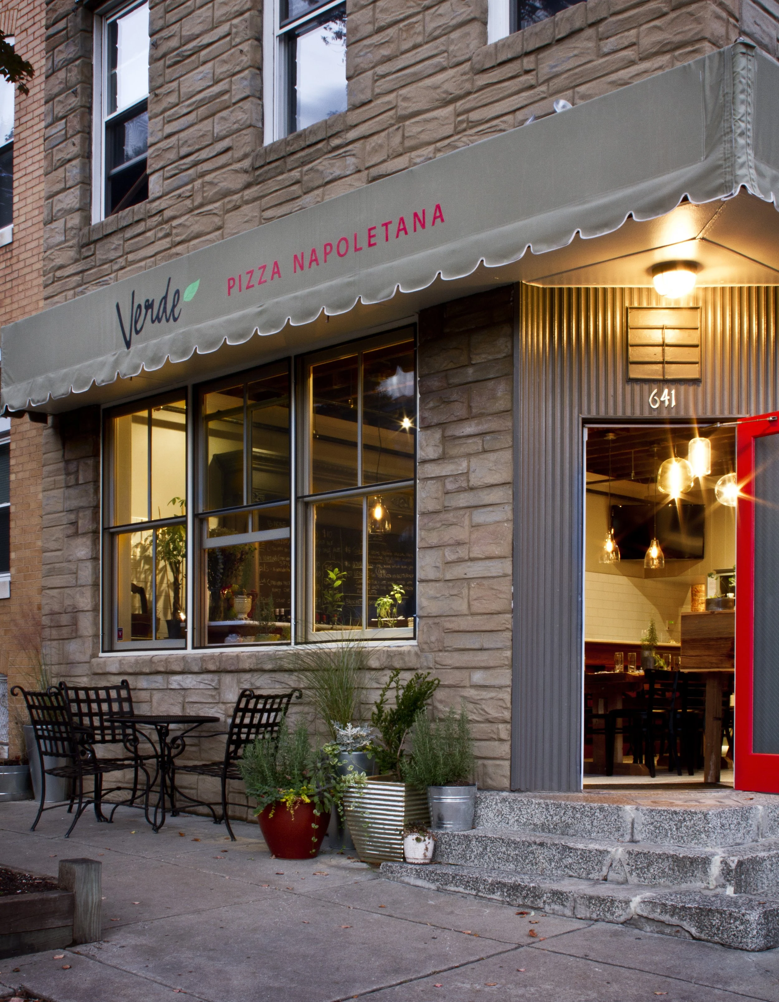 Exterior of a restaurant with a beige awning that has the words 'Verde Pizza Napoletana' written on it. The restaurant has large windows displaying the interior lighting and a cozy dining area. Outside, there are potted plants and metal chairs on the sidewalk. The building is made of brick with a stone façade around the entrance, which has the number 641 displayed above the door.