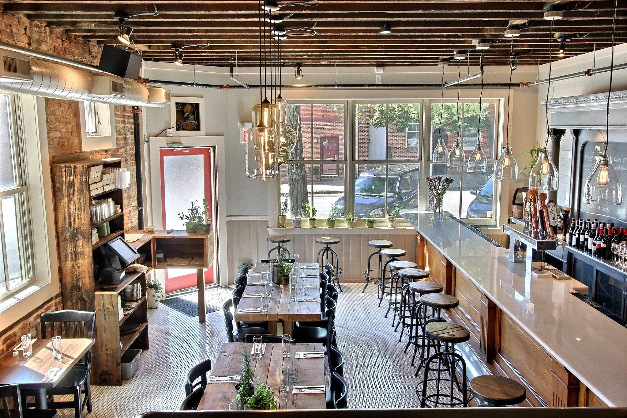 Interior of a modern restaurant or cafe with exposed brick walls, large windows, wooden furniture, pendant lighting, and bar seating. Plants and decorative elements are visible, with a view of parked cars outside.