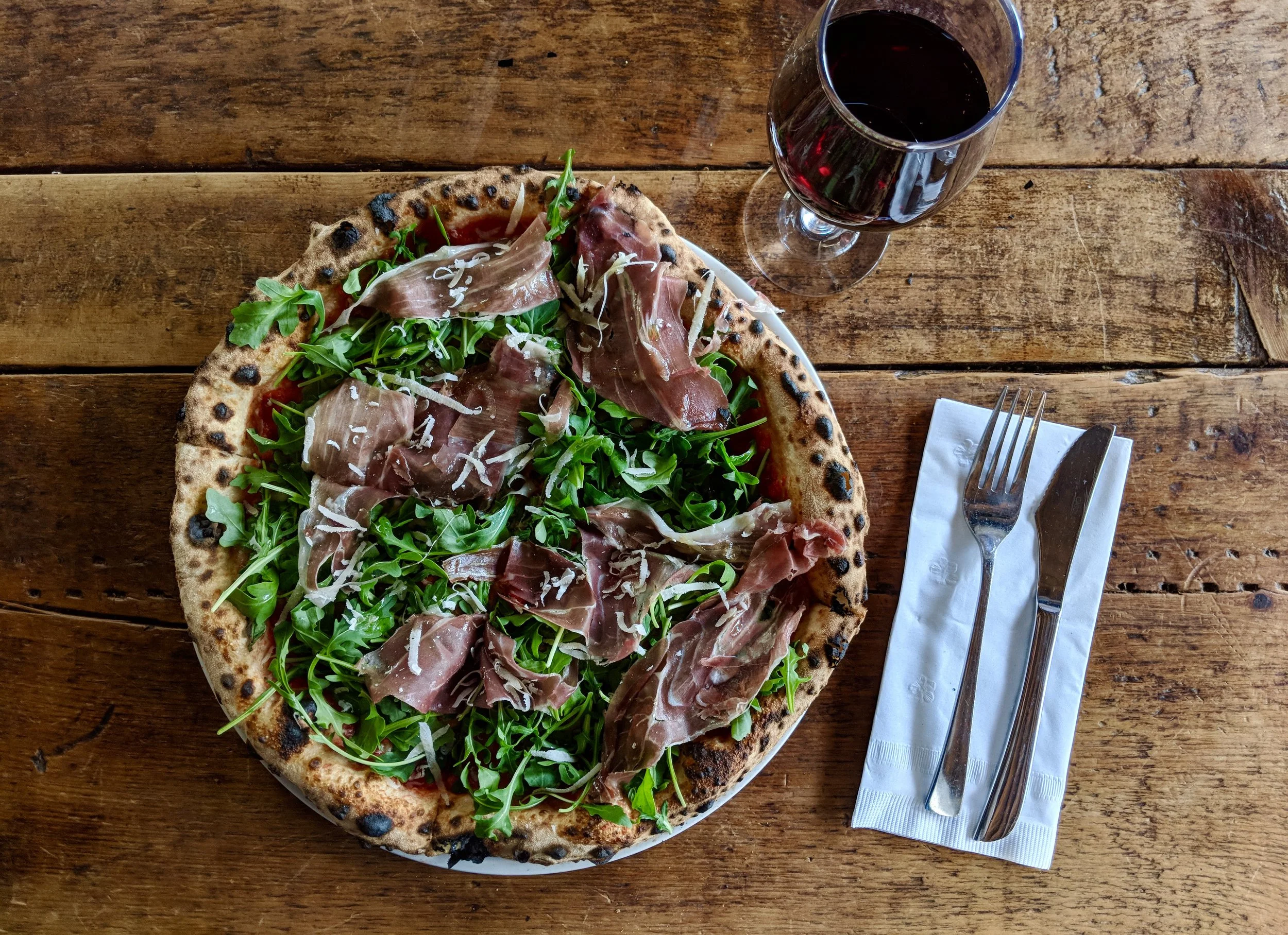 A pizza topped with prosciutto, arugula, and shredded cheese on a rustic wooden table, with a glass of red wine and a fork and knife on a napkin beside it.