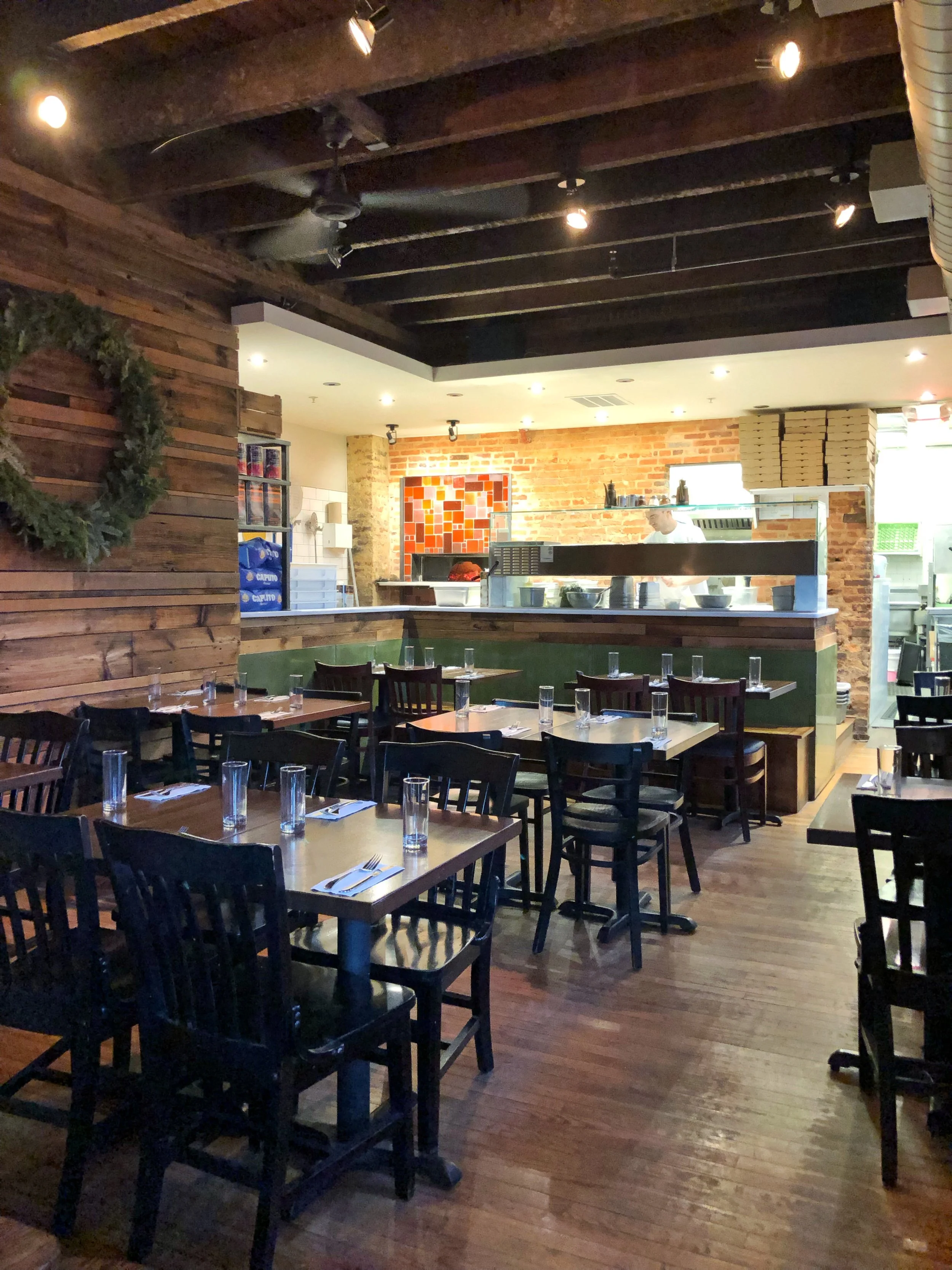 Inside a restaurant with wooden floors, tables set with glasses and utensils, and a bar area in the background with a brick wall and a kitchen staff member preparing food.