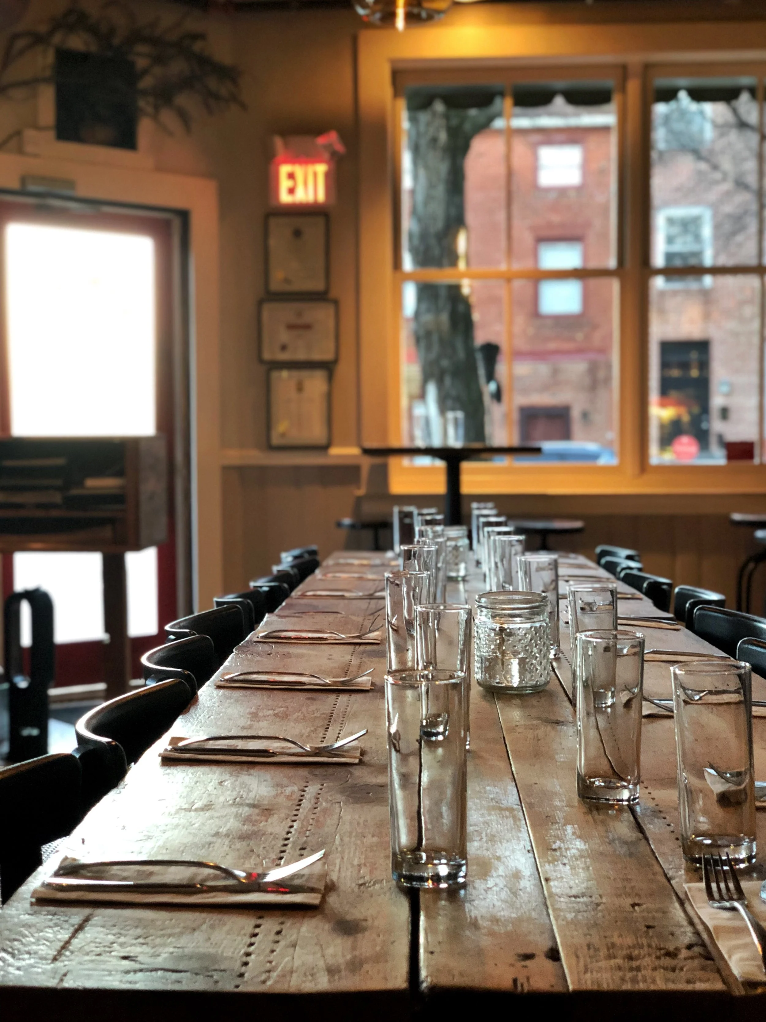 A long wooden dining table set with glasses, napkins, and utensils in a cozy restaurant with large windows and outside brick buildings.
