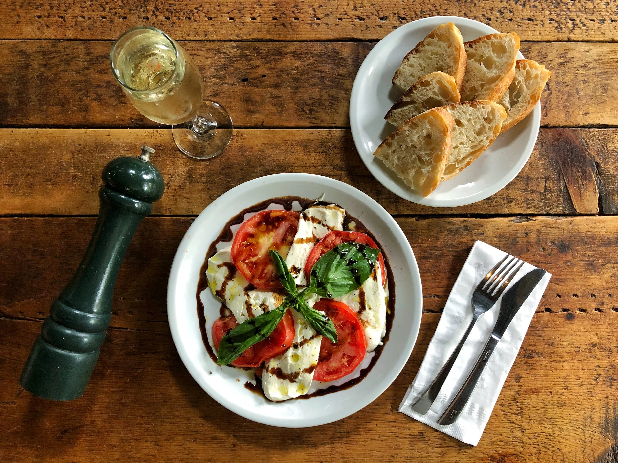 A plate of caprese salad with sliced tomatoes, fresh mozzarella, and basil leaves drizzled with balsamic glaze. Next to it are slices of focaccia bread on a white plate. There's a glass of white wine, a pepper grinder, and a fork and knife on a napkin on a wooden table.