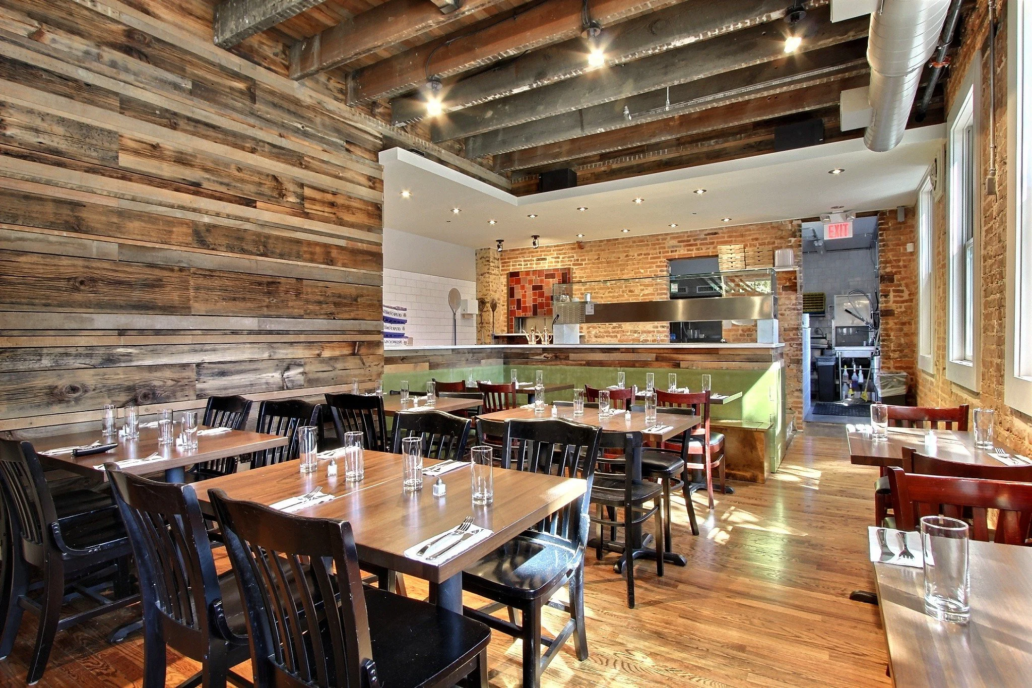 Interior of a restaurant with wooden tables and black chairs, set with glasses and utensils. Exposed brick walls and wooden ceiling beams are visible, with natural light coming through windows.